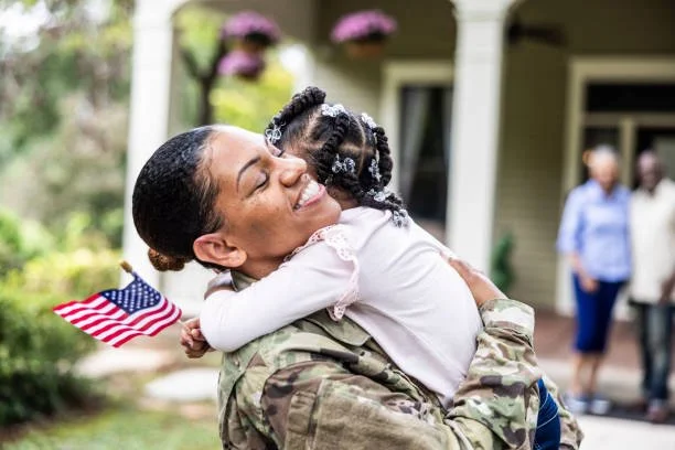 A woman in military uniform hugging a young girl, who is holding a small American flag, outdoors in a garden or yard.
