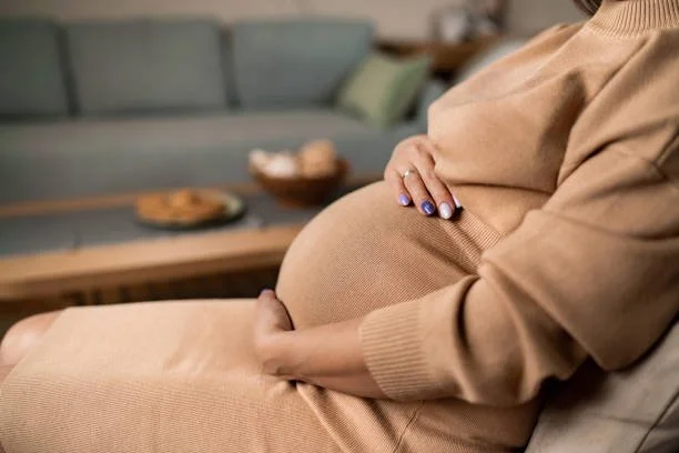 Close-up of a pregnant woman sitting on a couch with her hand on her belly, in a cozy living room setting.