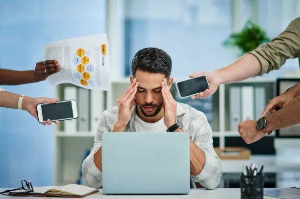 Man sitting at a desk with a laptop, surrounded by multiple people holding smartphones and papers, appearing stressed.
