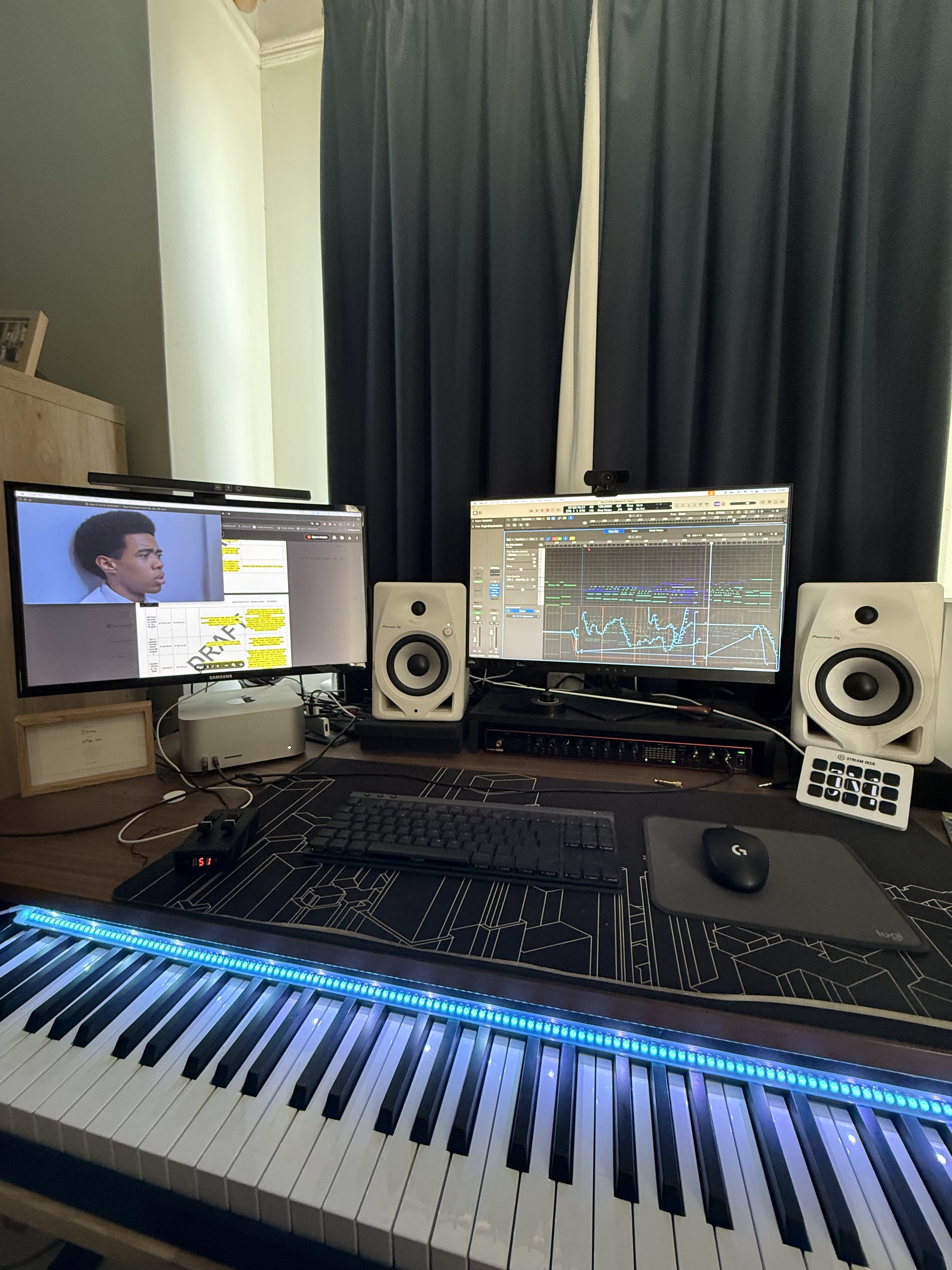 Home music production setup with a digital piano, dual computer monitors displaying audio editing software, studio speakers, a keyboard, mouse, and various electronic accessories, against a window with dark curtains.