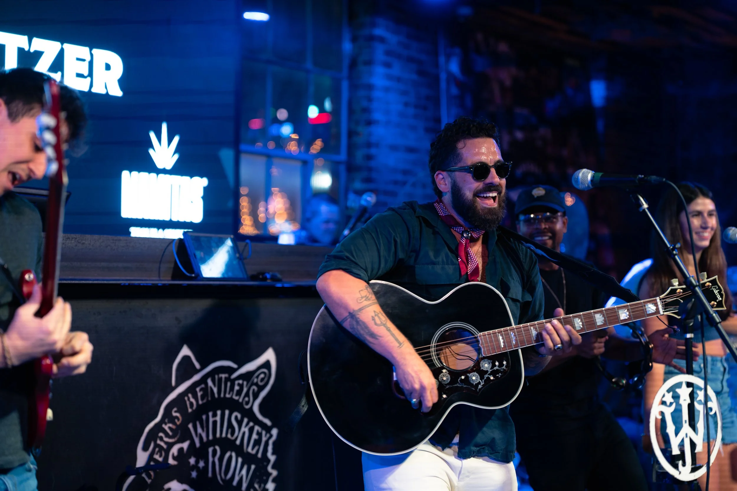 Omer Netzer has is performing music on stage at Whiskey Jam, with a man playing an acoustic guitar and singing, wearing sunglasses and a red bandana.