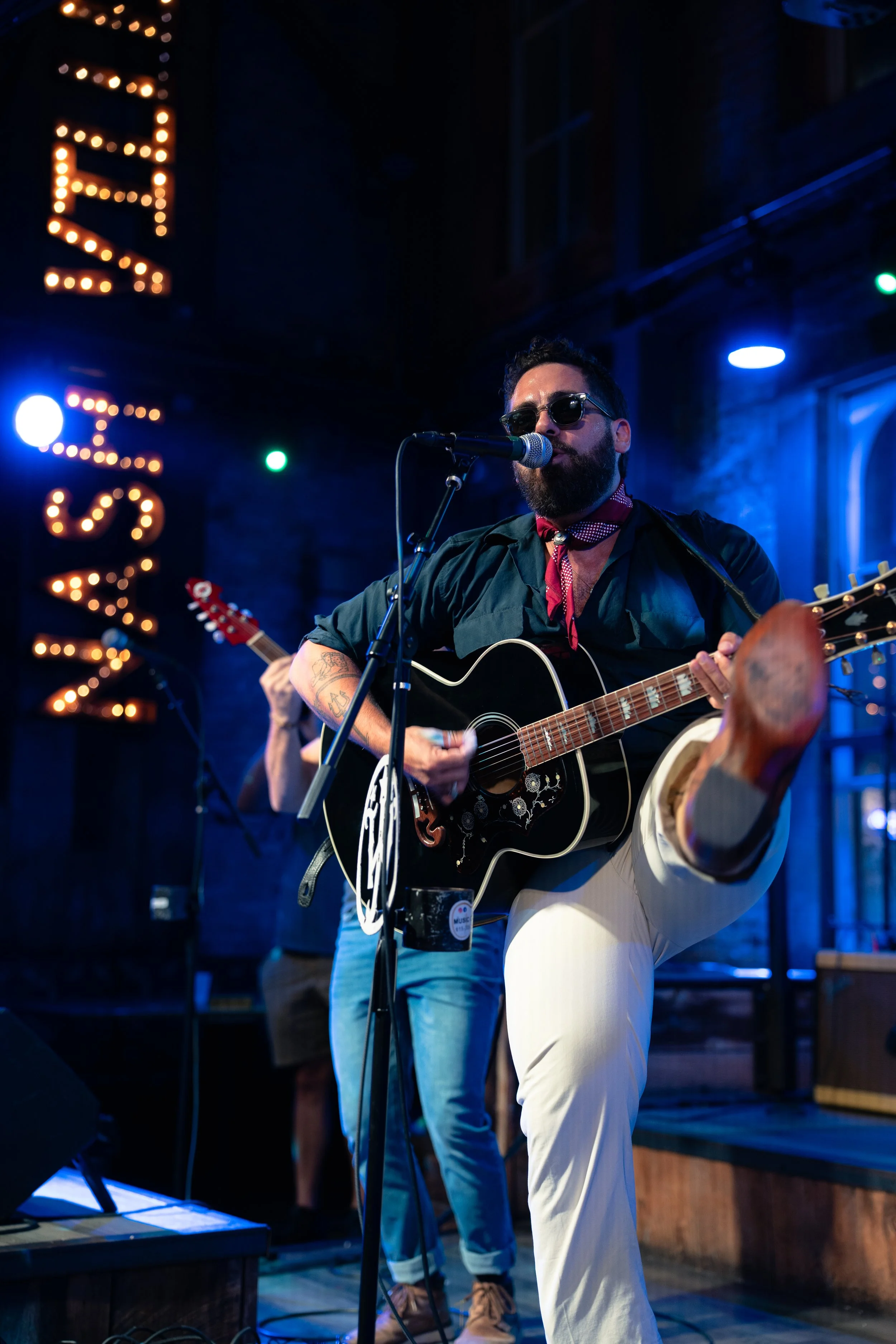Omer Netzer is playing an acoustic guitar on stage during a live music performance at Whiskey Jam, with a colorful neon sign that says 'MUSIC' in the background.