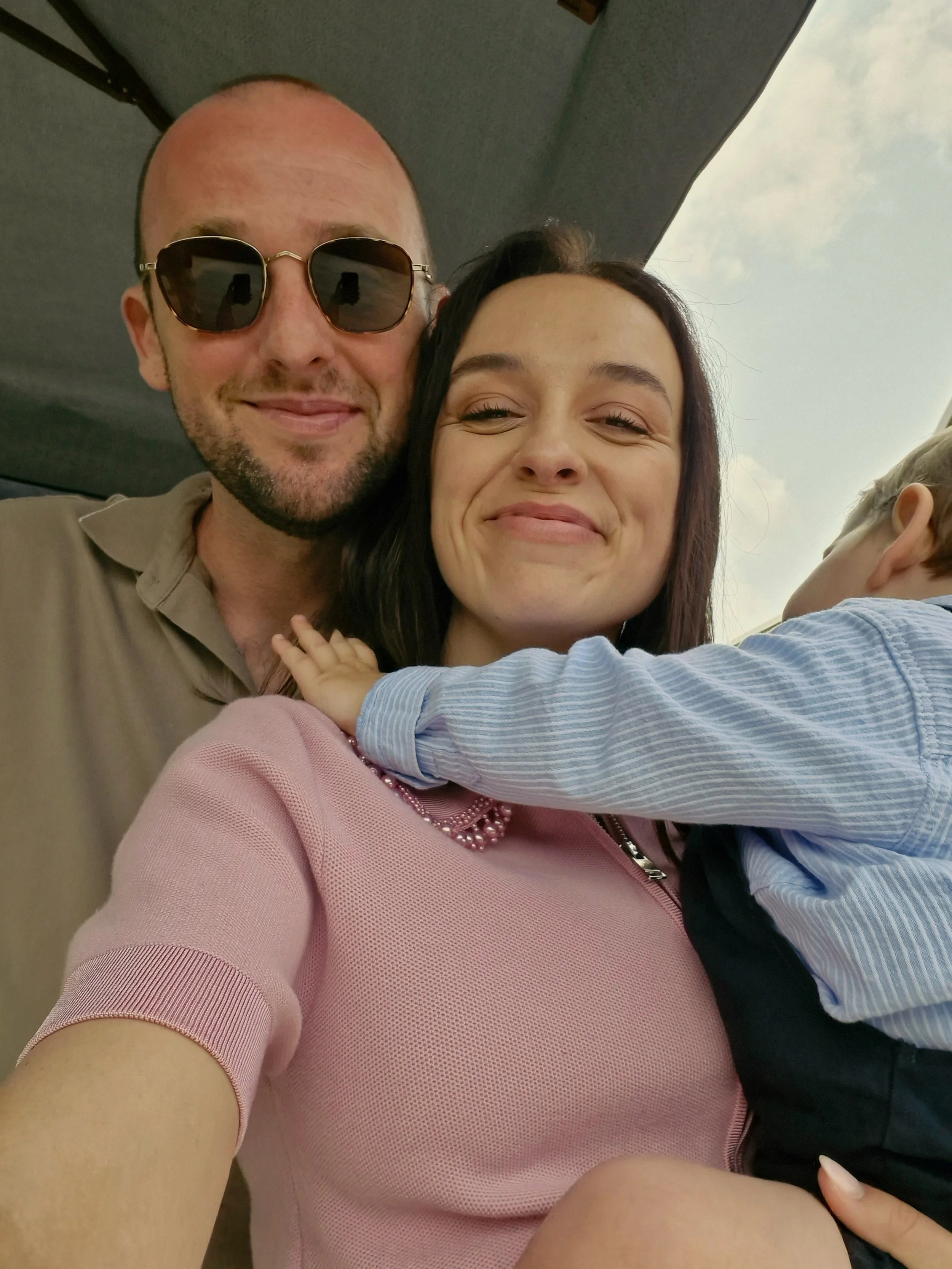 Drie mensen die een selfie nemen, een man met zonnebril, een vrouw en een kind dat in haar armen ligt, met een blauwe lucht op de achtergrond.