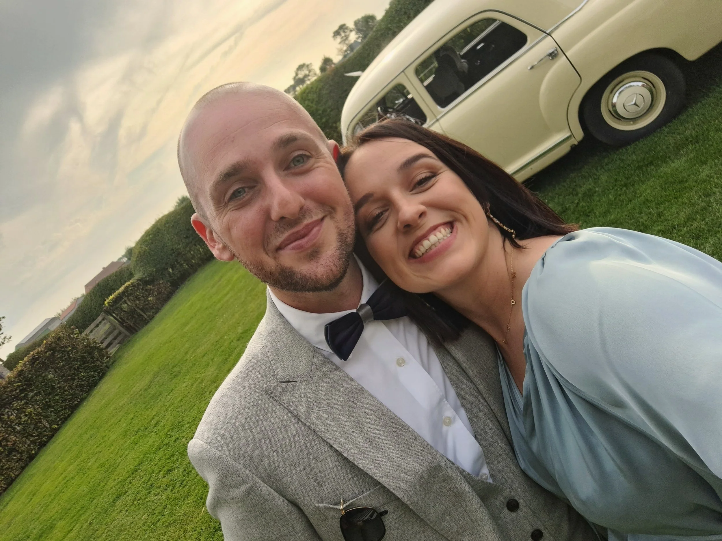 Man en vrouw knuffelen en glimlachen in een groene tuin met een witte oldtimer auto op de achtergrond.