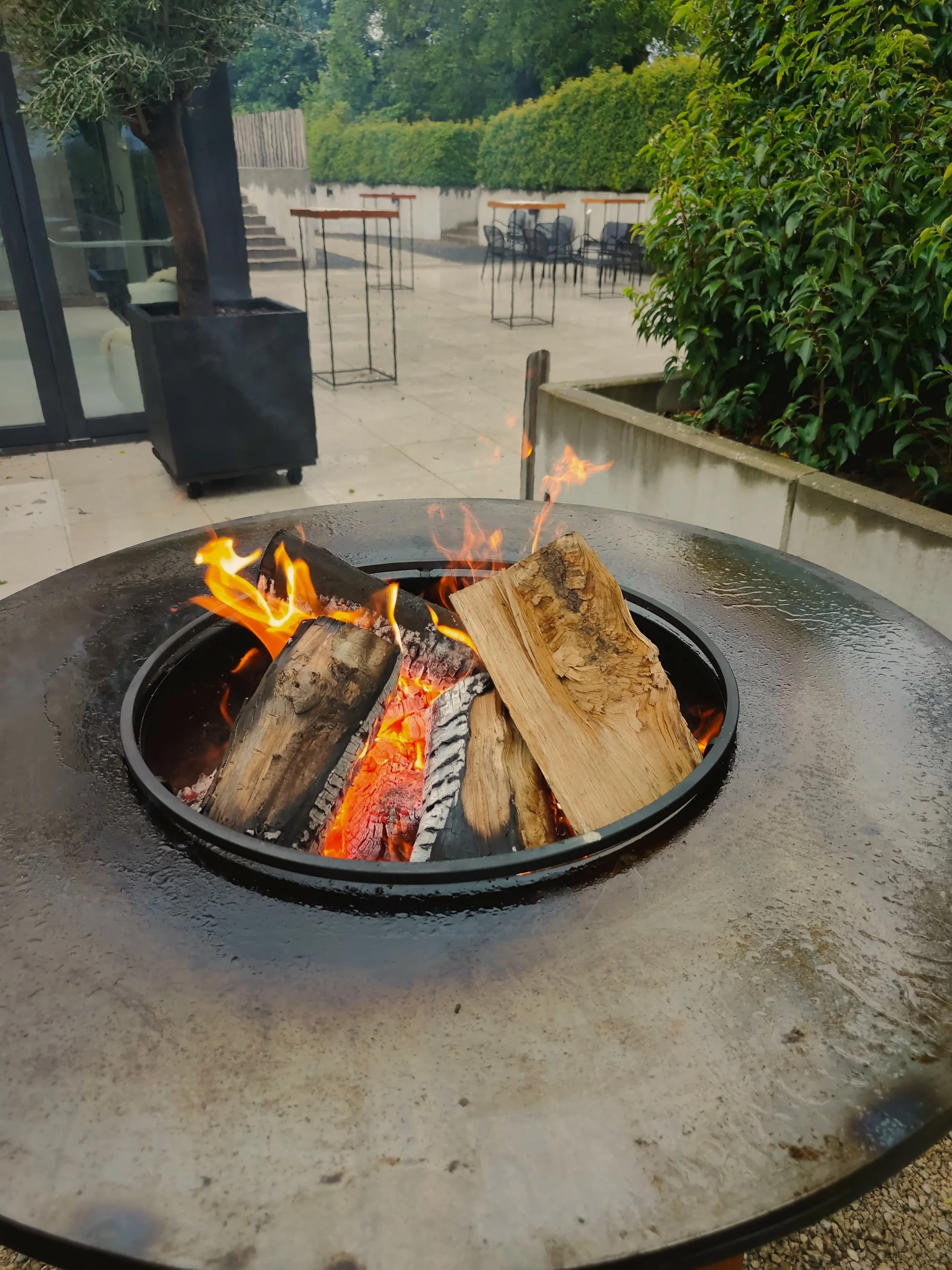Een vuurkorf met brandend hout op een buitentafel, met een tuin en patio meubilair op de achtergrond.