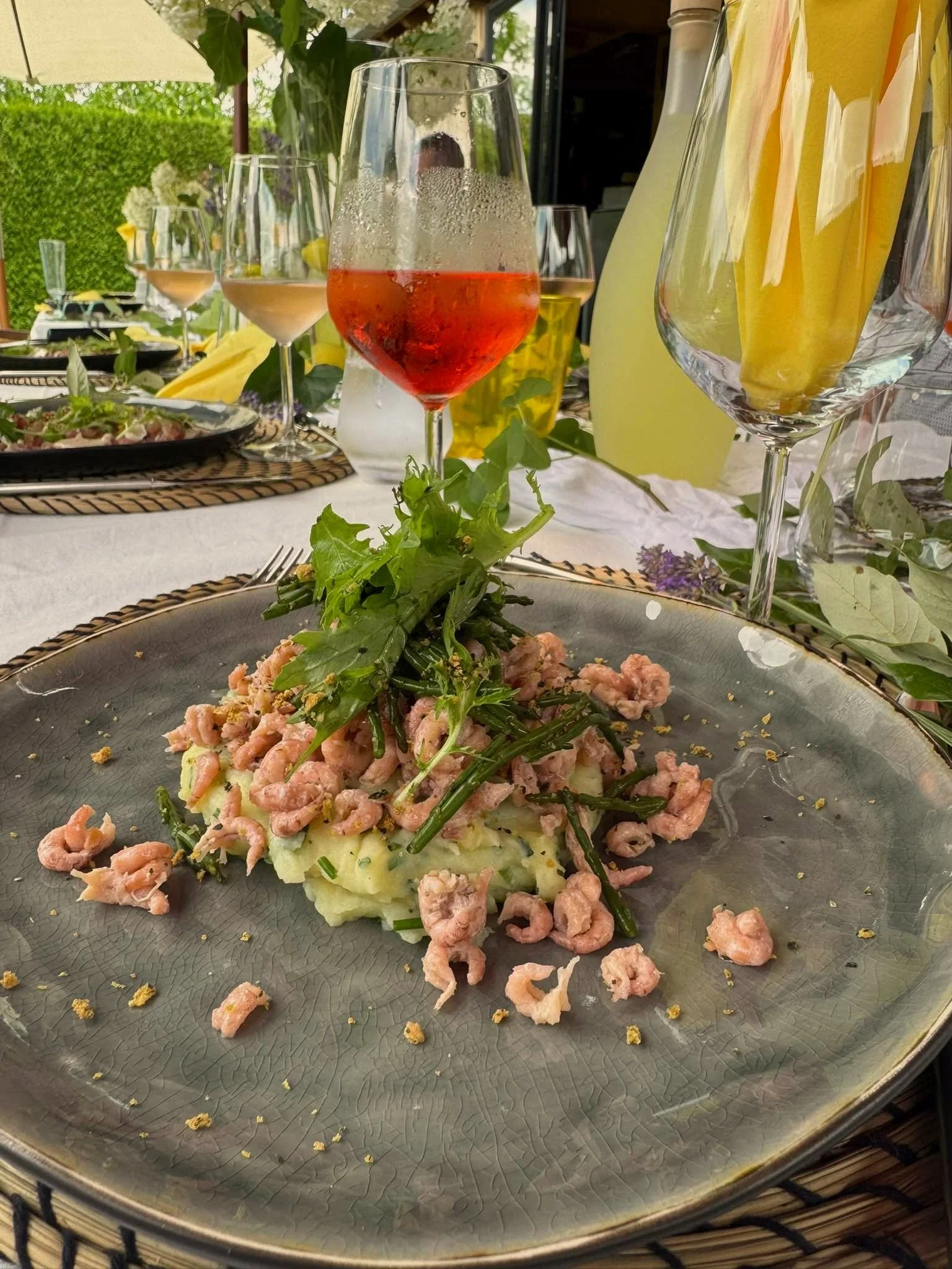 Gereserveerde tafel met een bord met garnalen, aardappelpuree en groene groenten, omgeven door verschillende glazen met wit, rood en rosé wijn, en een achtergrond van bloemstukken en tuin.