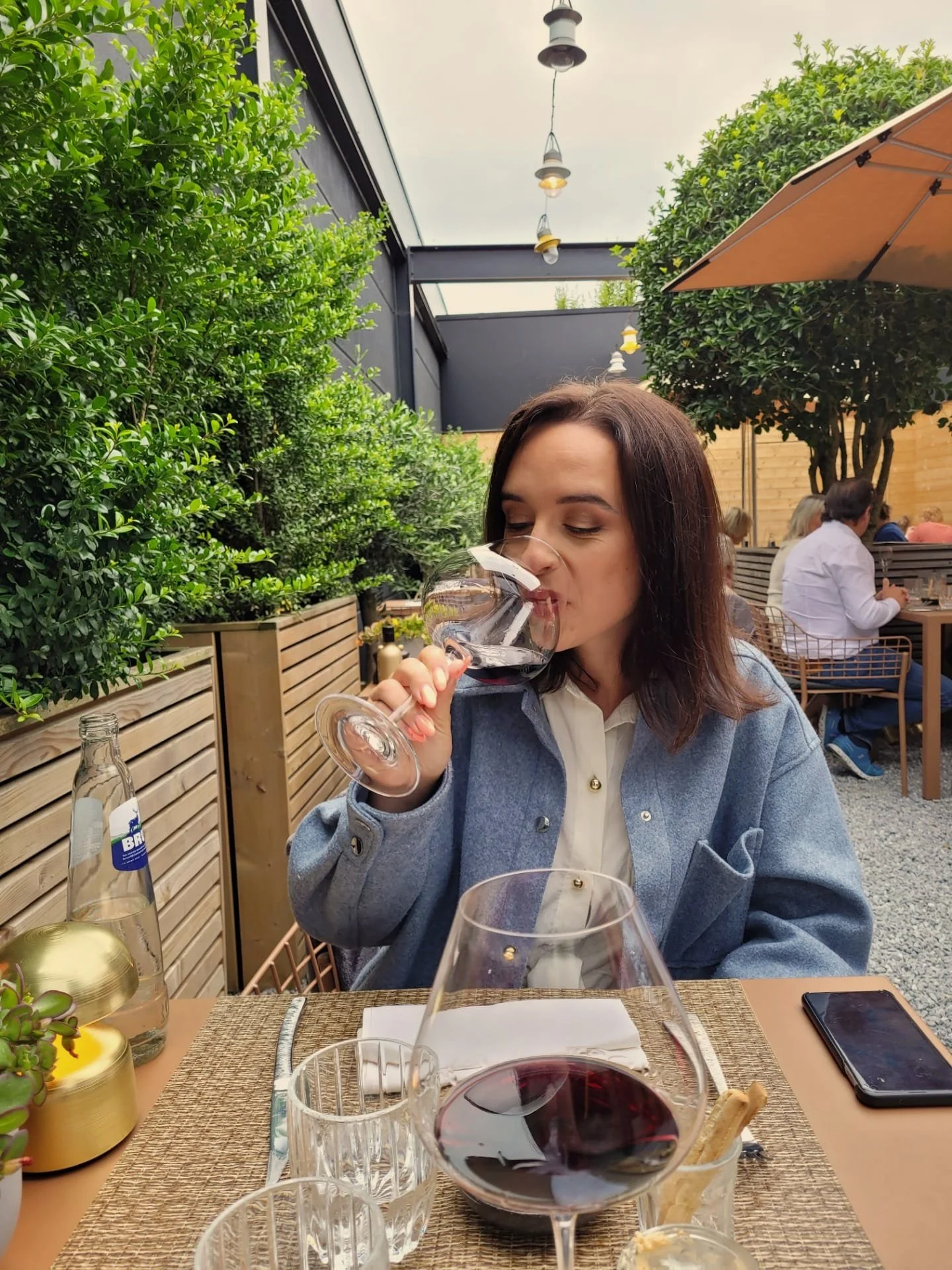 Vrouw drinkt rode wijn in een restaurant met groene planten, houten omheining, en andere gasten op de achtergrond.