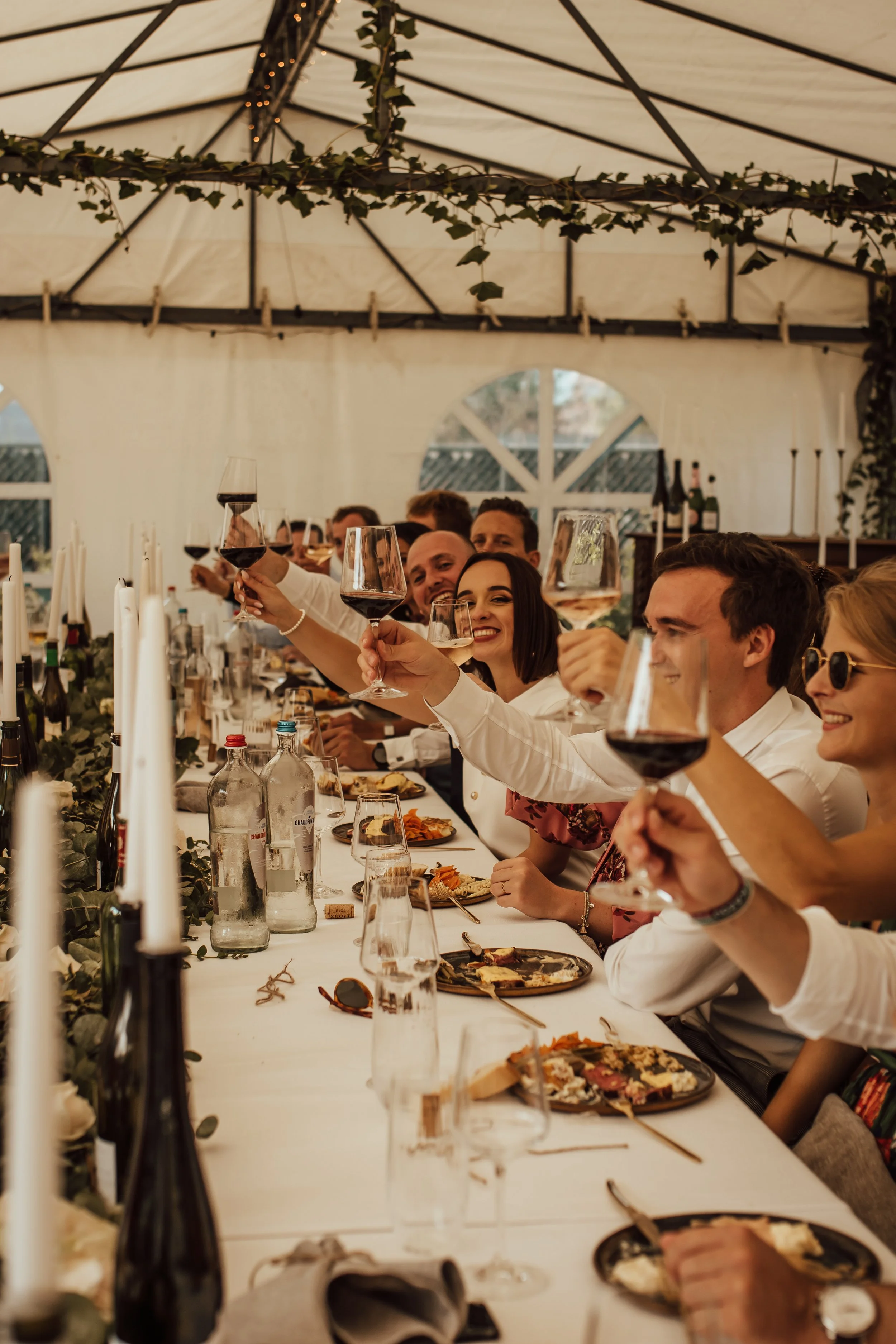 Mensen zitten aan een lange tafel in een tent, proosten met glazen rode wijn tijdens een feestelijk diner.
