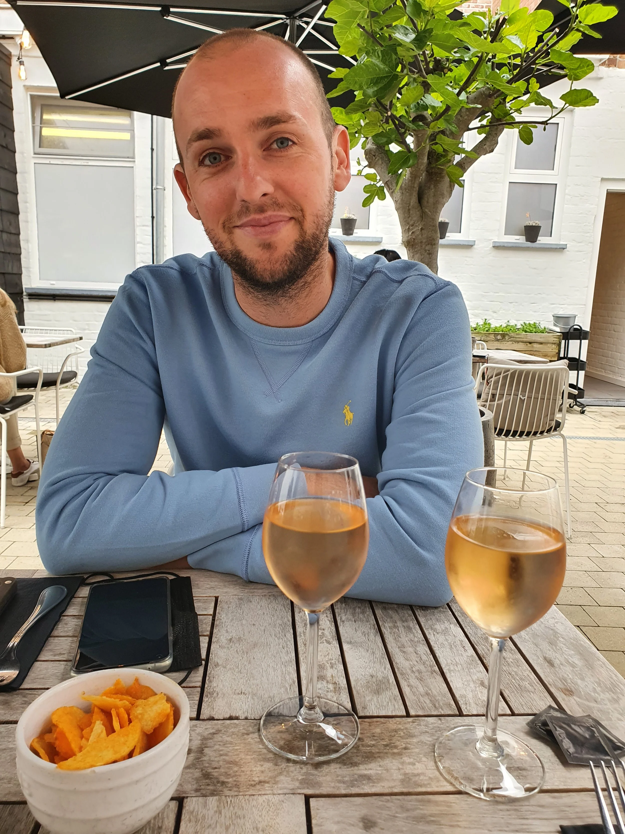 Man zittend aan een houten tafel met twee glazen witte wijn, een kom met chips, en een telefoon, buiten op een terras met planten en witte muren.