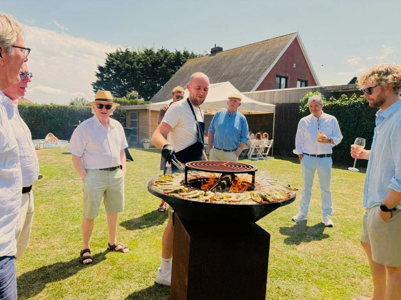 Mensen genieten van een barbecue in een tuin op een zonnige dag, met een man die eten op een ronde grill bereidt terwijl anderen lachen en drinken.
