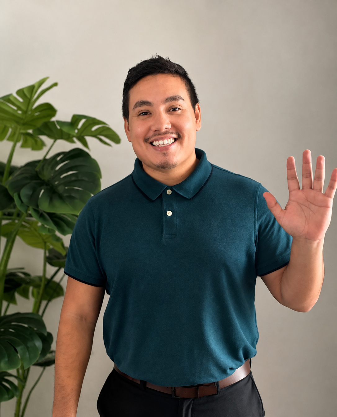A smiling man with short dark hair and a mustache, wearing a teal polo shirt, waving with his right hand, standing next to a large potted monstera plant against a plain white wall.