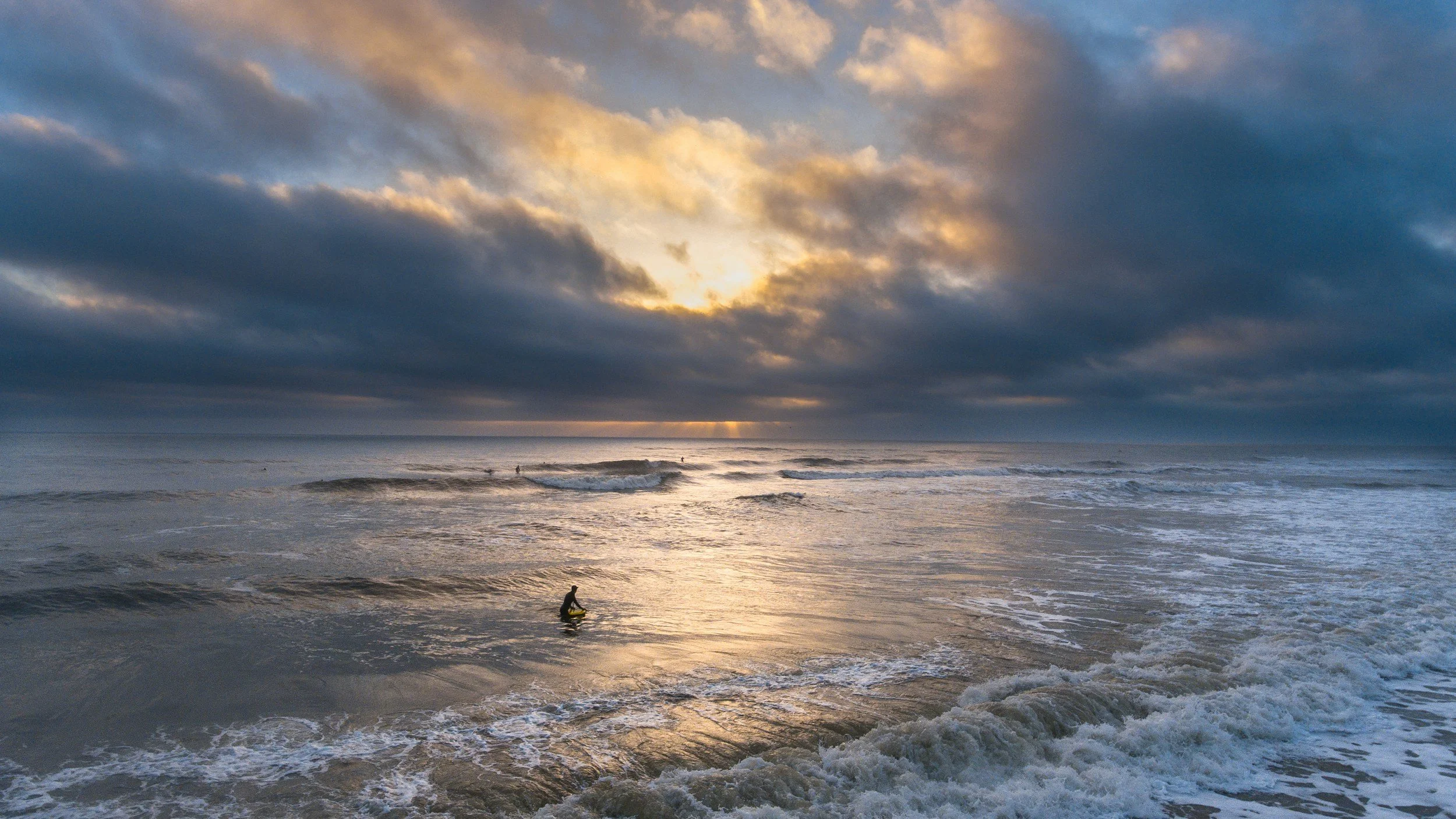 Sunset over the ocean with dark clouds, small waves, and a lone surfer in the water.