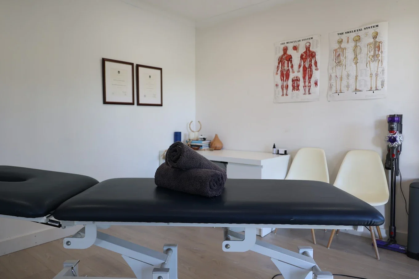 Physical therapy or chiropractic treatment room with a black treatment table, two rolled black towels, framed certificates on the wall, anatomical posters, white chairs, and therapy equipment in the background.