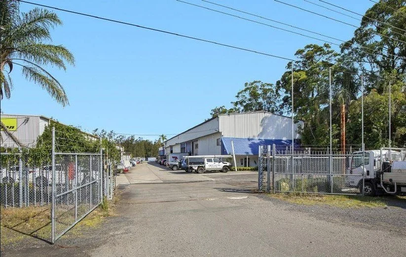 A view of a warehouse or industrial building with a fenced parking lot, several trucks, and trees in the background under a clear blue sky.