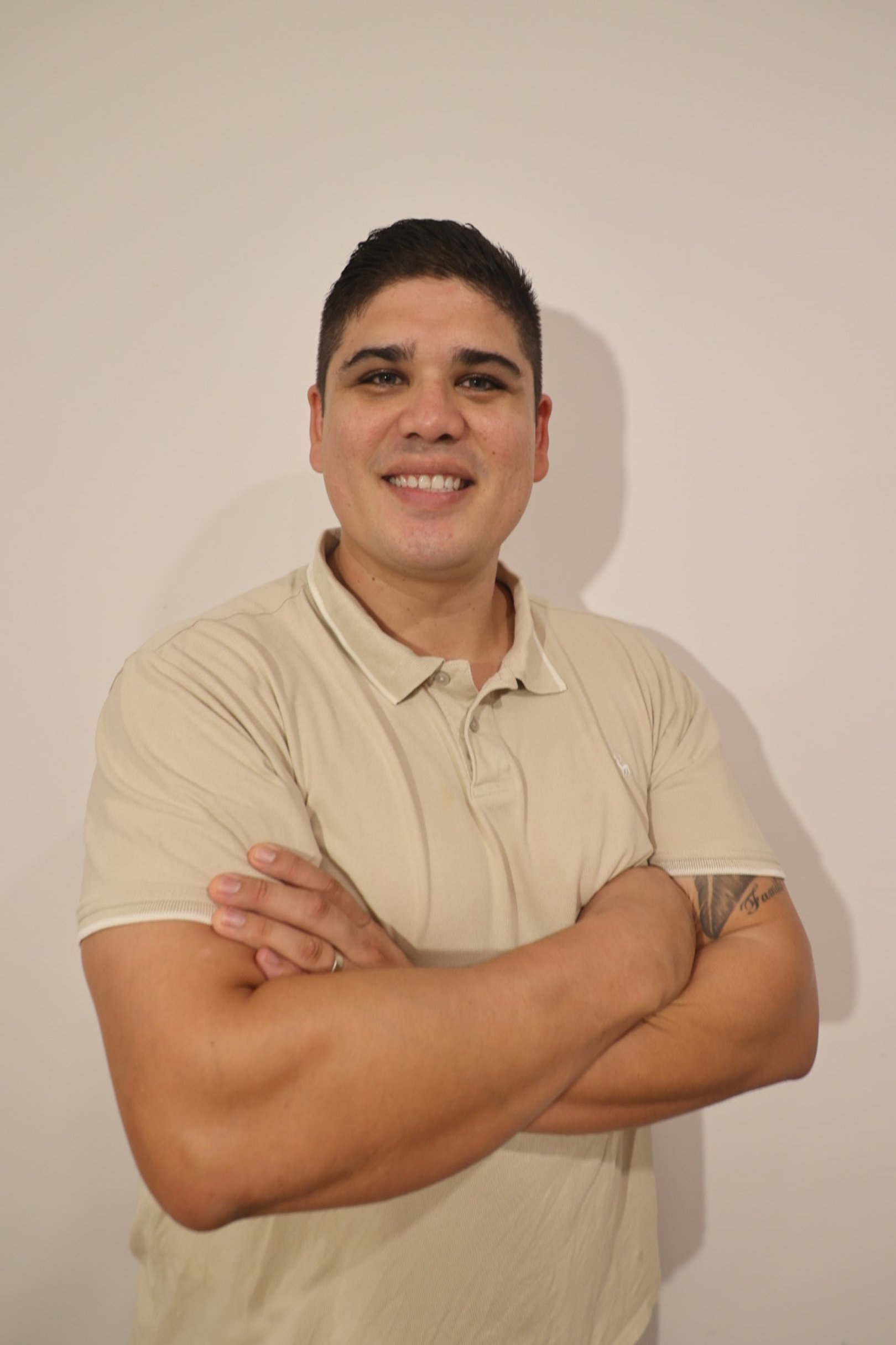 A young man with short dark hair smiling and crossing his arms, wearing a beige polo shirt, standing against a plain white wall.