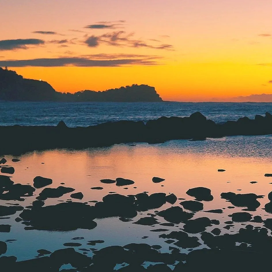 A sunset over the ocean with rocks in the foreground and silhouetted landforms in the distance.