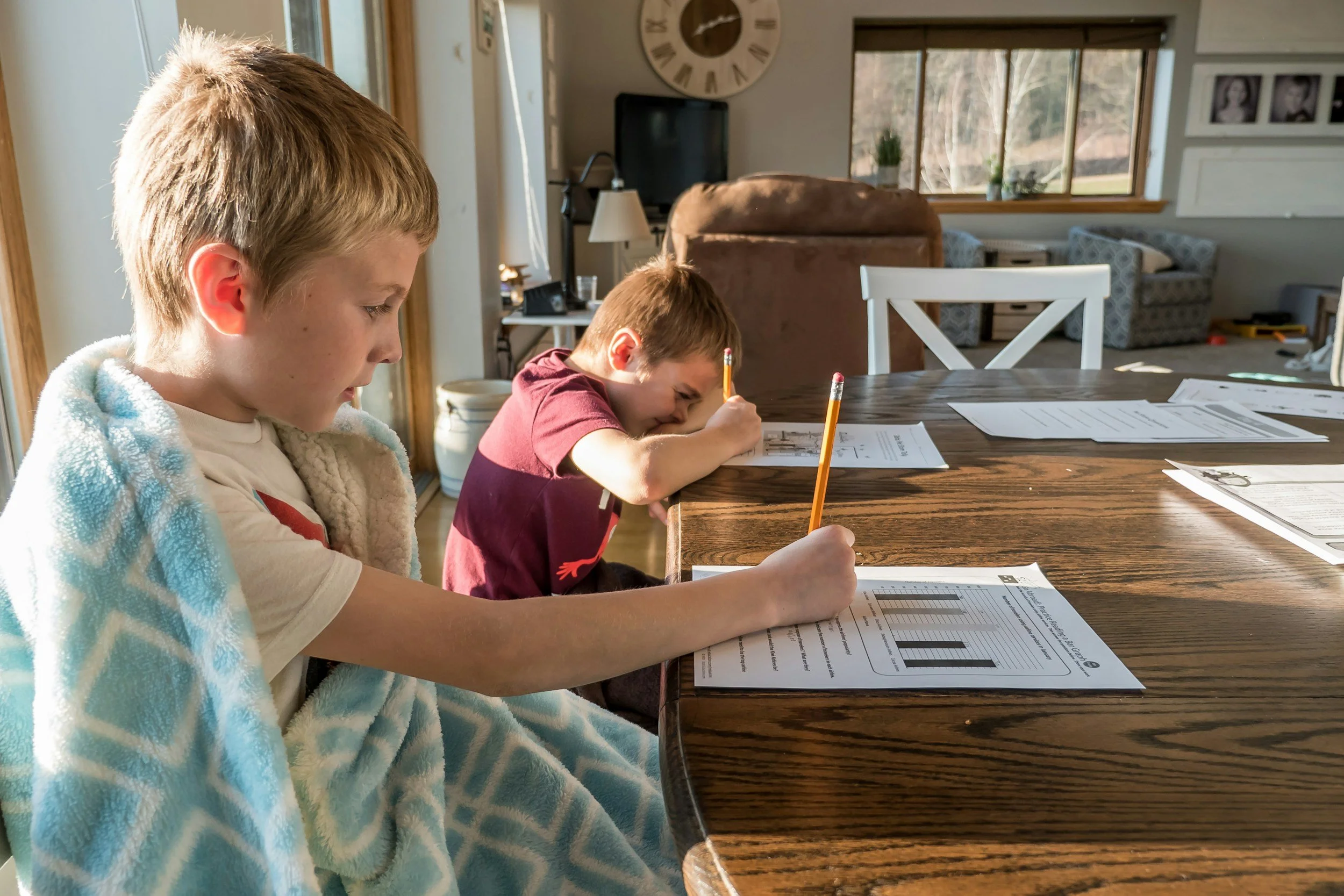 Two young boys sit at a wooden table working on school worksheets, one with a blanket over his shoulders. They are inside a bright kitchen or dining area with large windows, a wall clock, and framed photos visible in the background.