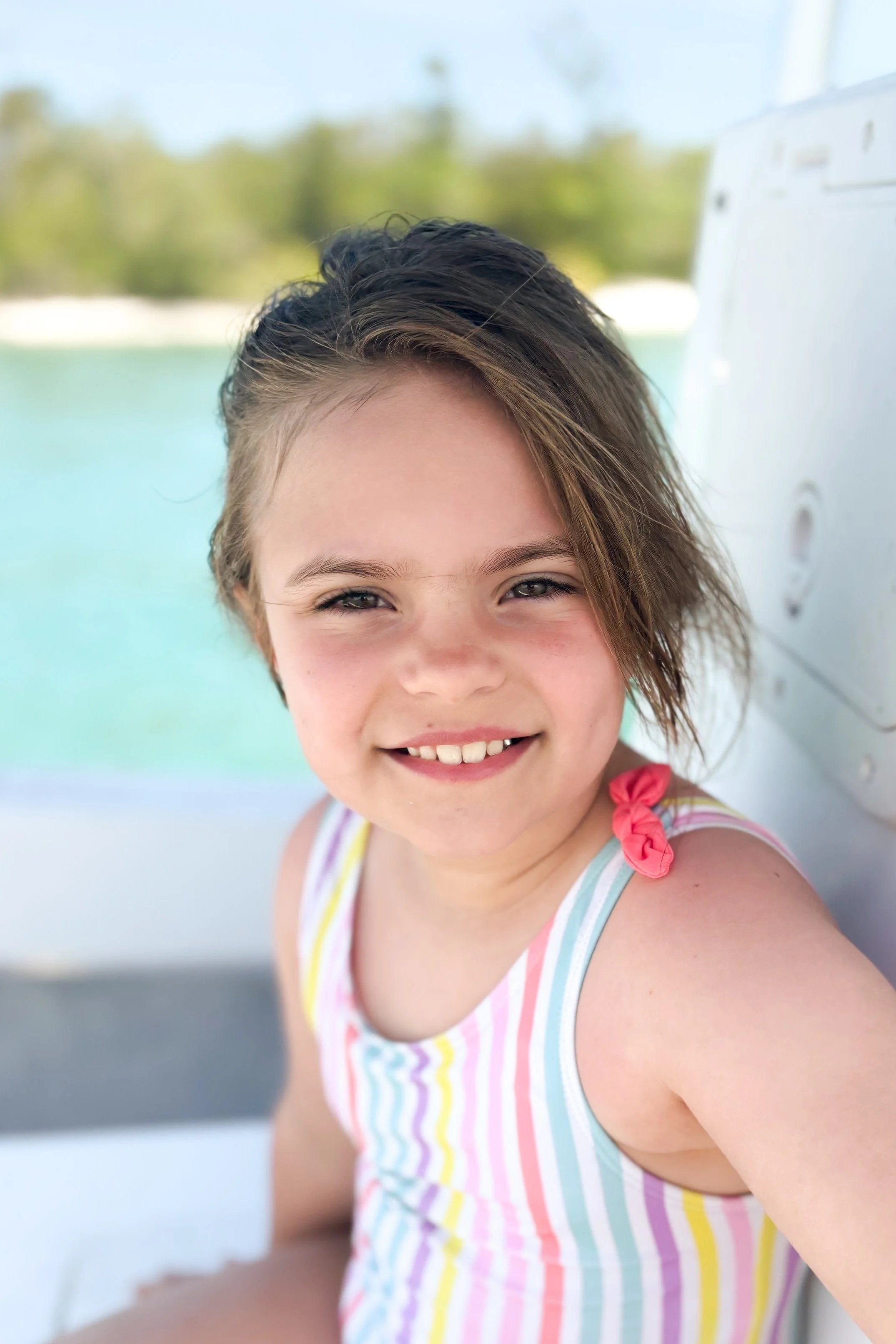 A smiling girl with short brown hair, wearing a colorful striped tank top with pink, yellow, blue, and purple colors, near a body of water.