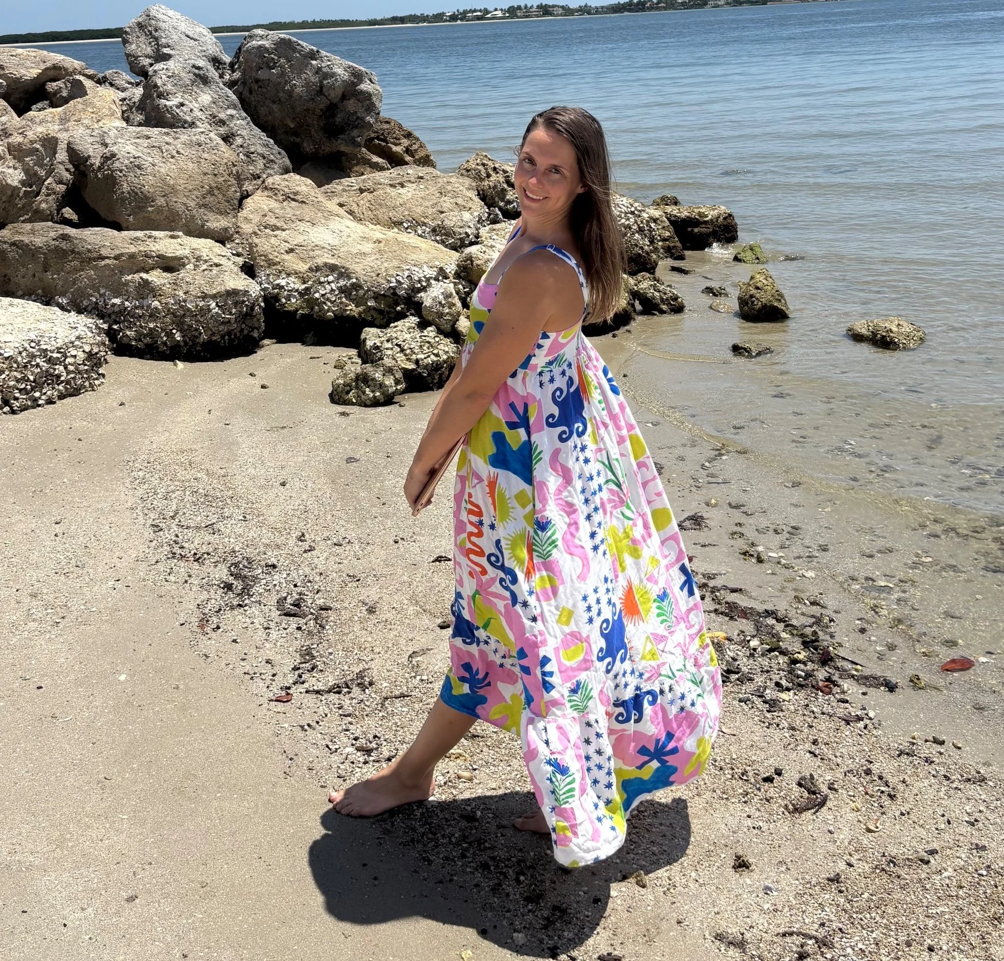 A woman standing on a sandy beach near water with rocks in the background. She is smiling, wearing a colorful, patterned dress, and appears barefoot. The weather is sunny.