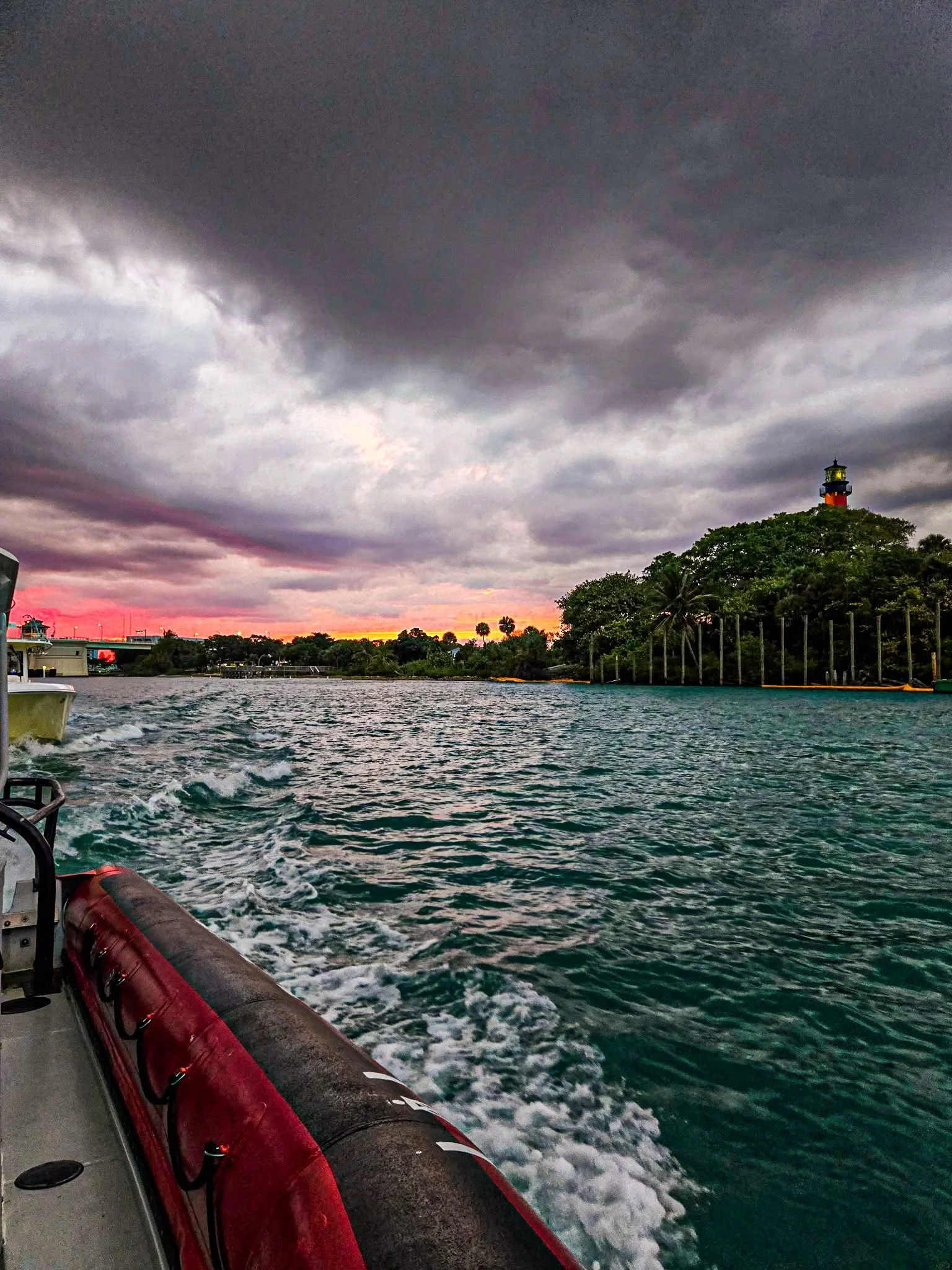 The photo shows a view from a boat on the water, with a red buoy or life raft on the left side. The water is choppy, and the sky above is cloudy with dark storm clouds, with a hint of a colorful sunset or sunrise near the horizon. In the background, there is a small hill or island with a lighthouse on top, surrounded by trees.