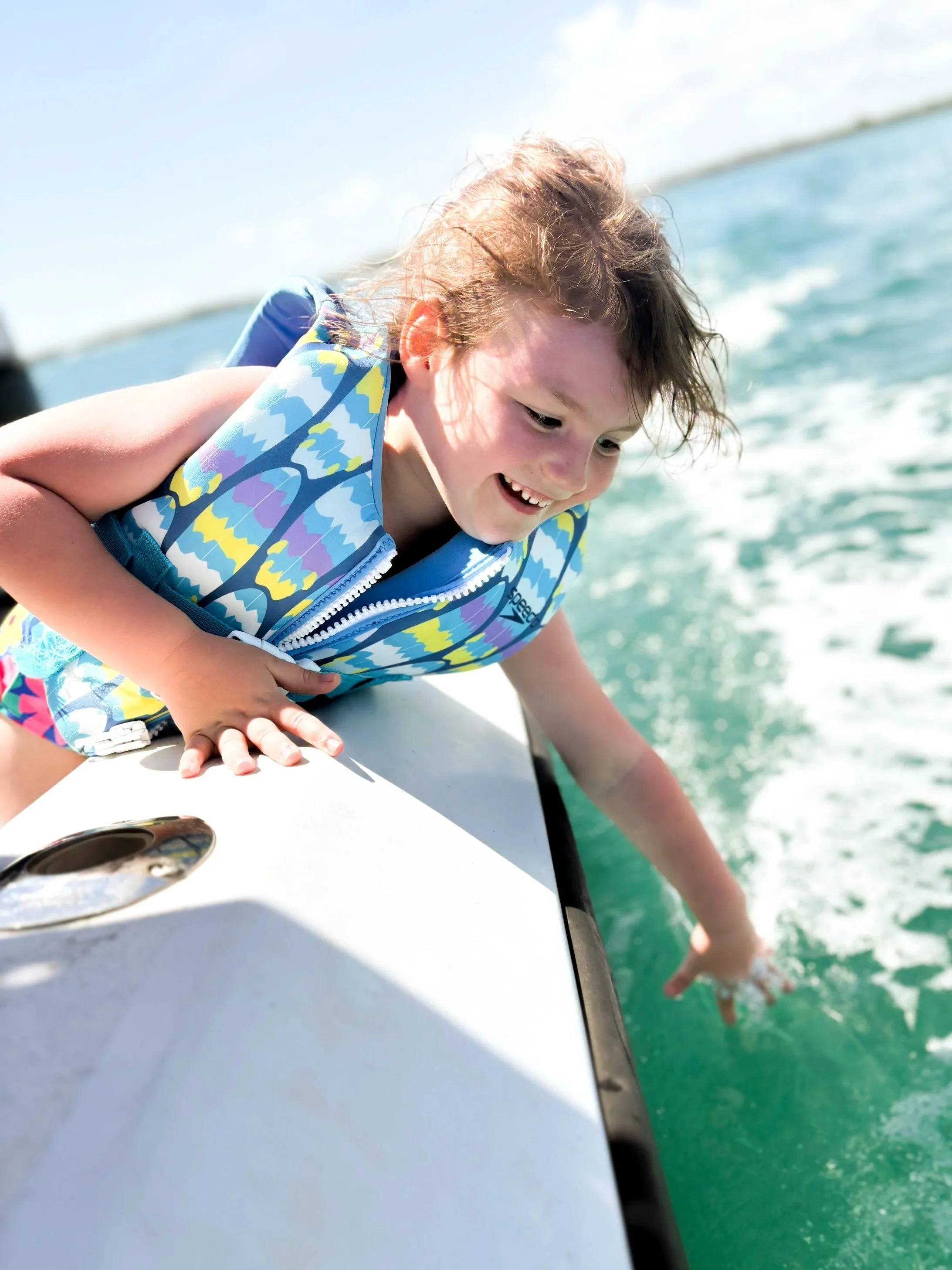 Child in life jacket on boat pointing into the water with a finger, holding onto the boat's edge, with background of water and sky.