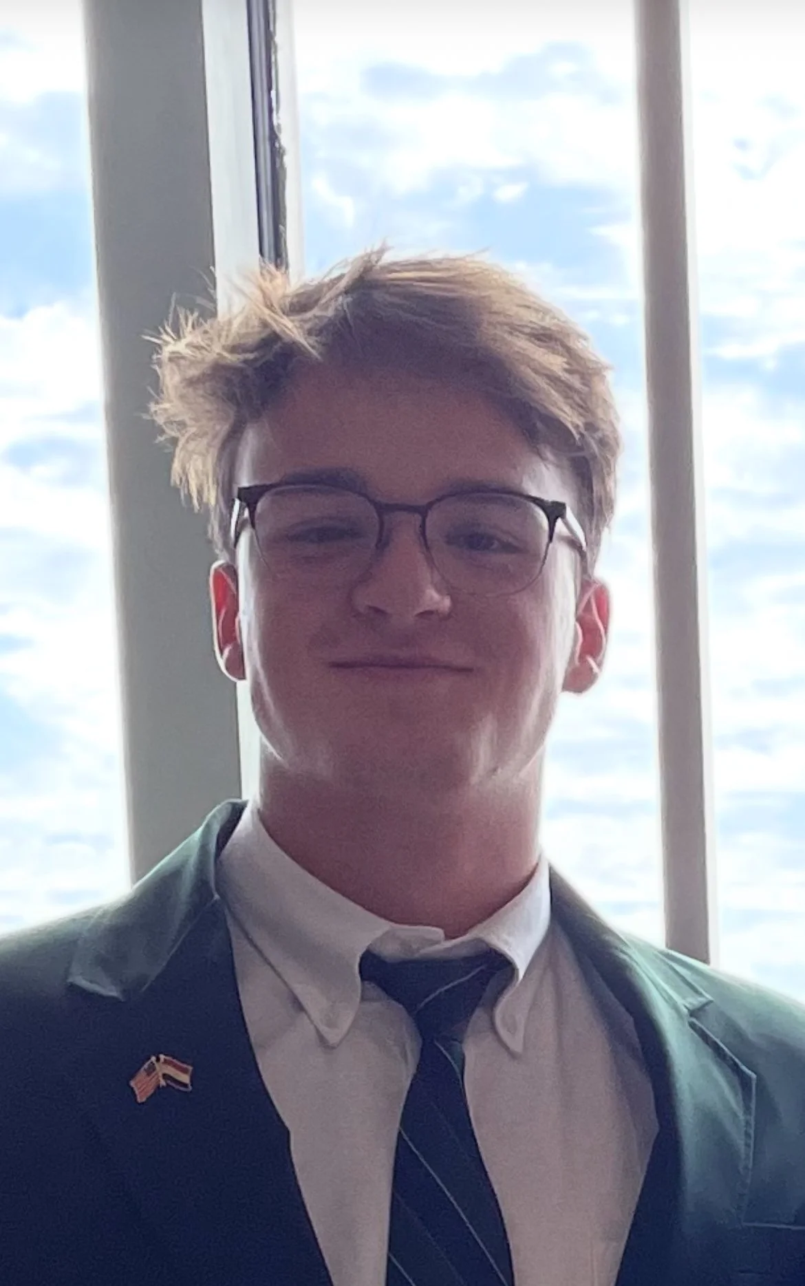 Young man wearing glasses, a white shirt, a striped tie, and a blazer with a small flag pin, standing in front of a window with a view of the cloudy sky.
