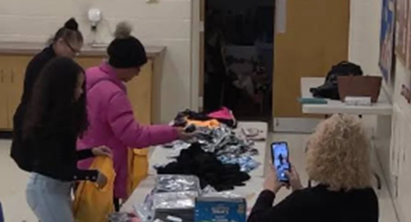 Three girls are shopping for clothes at a table with folded garments, while a woman with curly blonde hair is taking a photo with a smartphone.