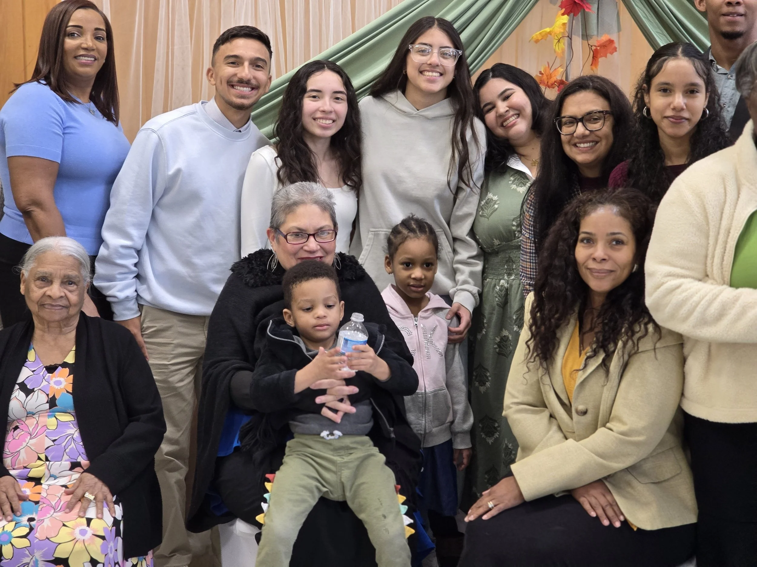 Group of diverse people, including children and adults, smiling and posing together indoors with autumn-themed decorations in the background.