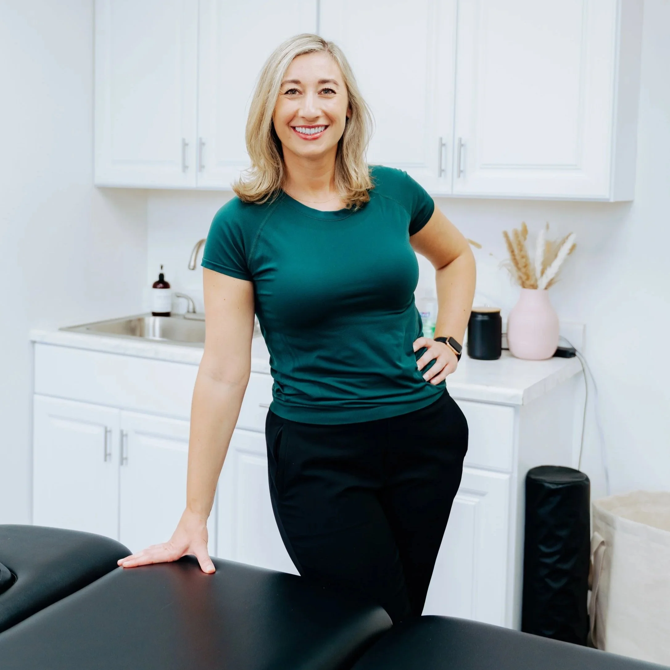 A woman with blonde hair smiling and wearing a teal shirt, standing in a bright, modern kitchen with white cabinets, a sink, and a pink vase with dried plants on the counter.