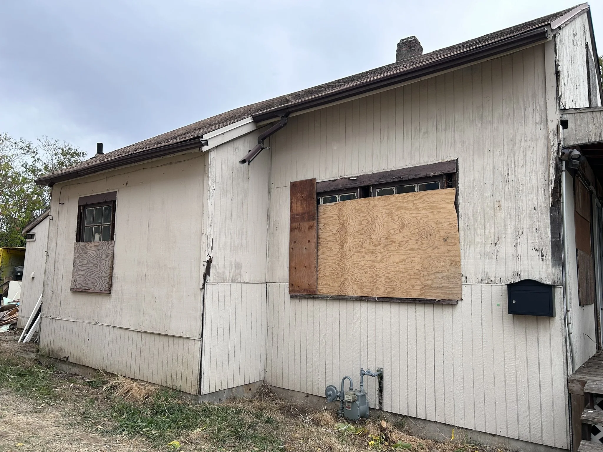 An old, weathered house with plywood covering the windows and an opening, showing visible damage and missing parts of the siding.