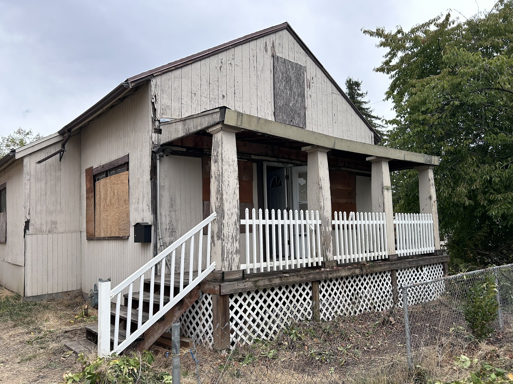 A house with boarded-up windows and a weathered exterior, featuring a small porch with a white picket fence and a damaged set of stairs.