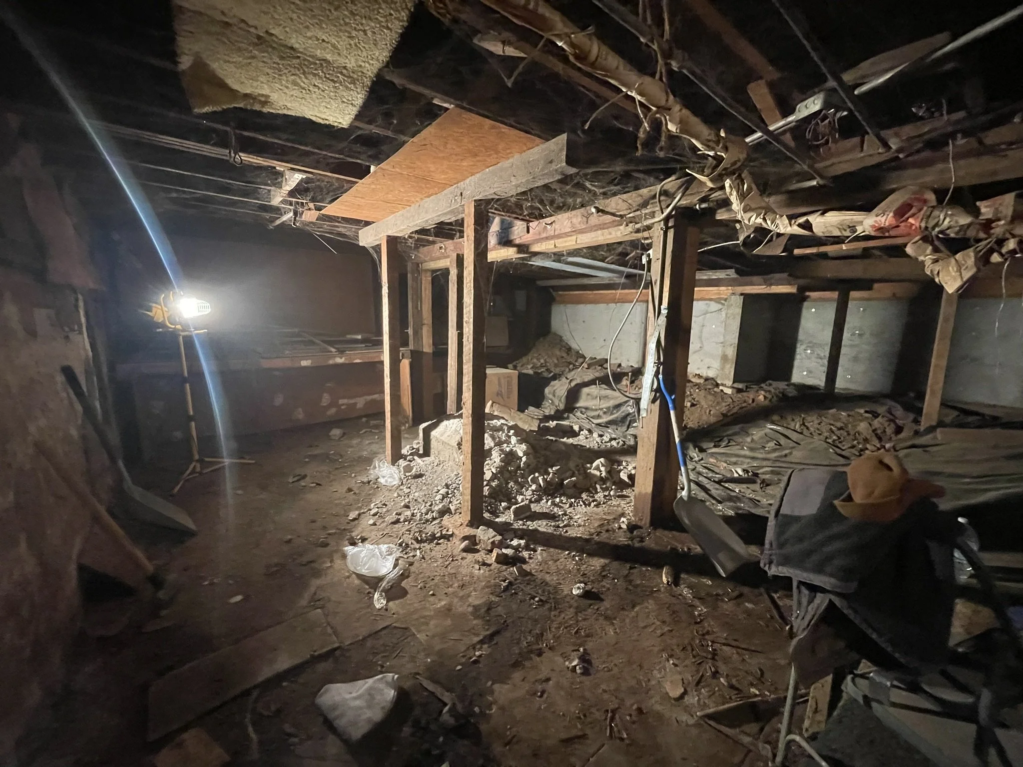 The image shows an unfinished basement with exposed wooden beams, electrical wiring, scattered debris, and dirt flooring. A bright work light is turned on, illuminating part of the space.