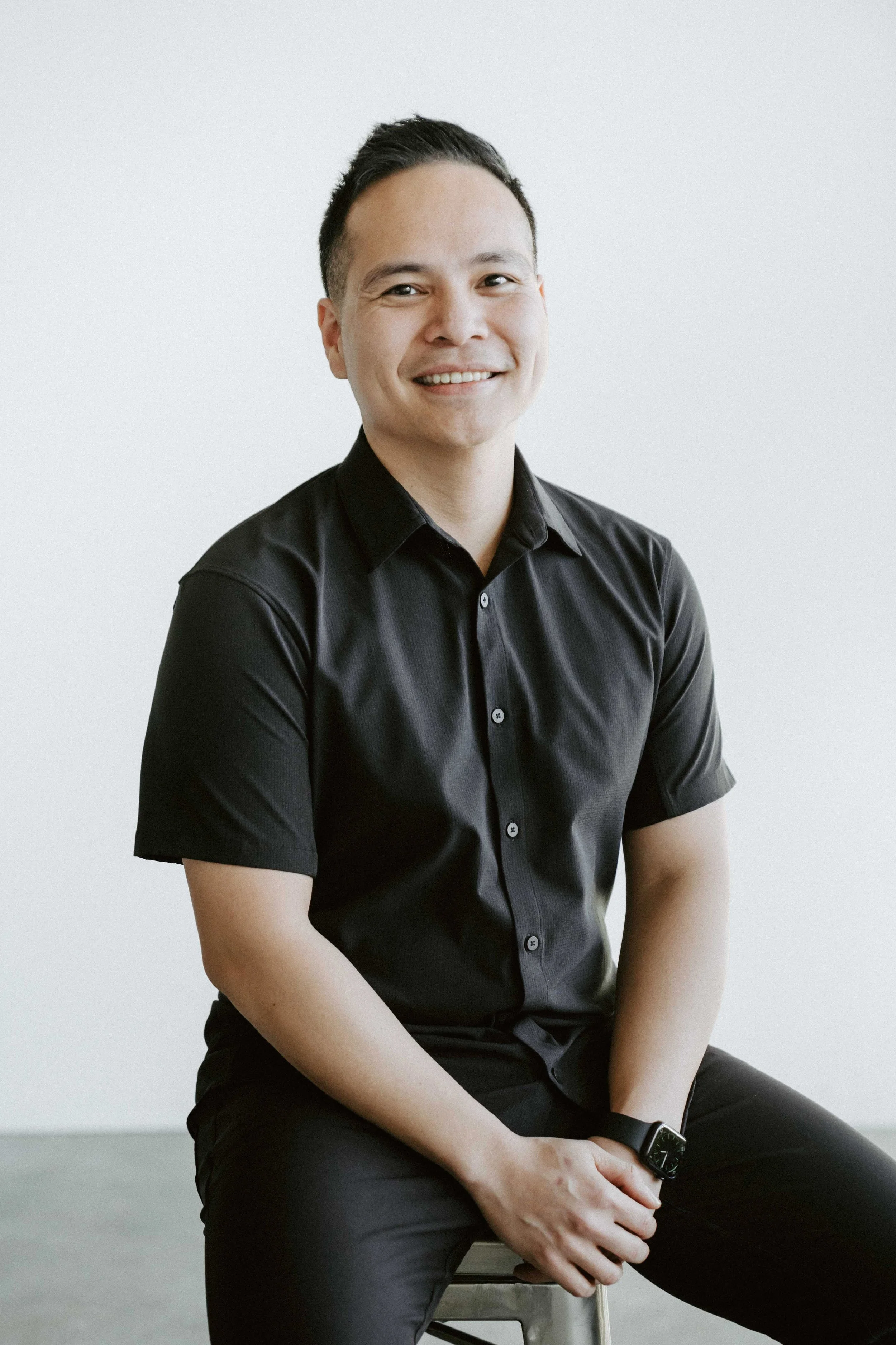 A smiling man in a black button-up shirt sitting on a stool against a plain light background.