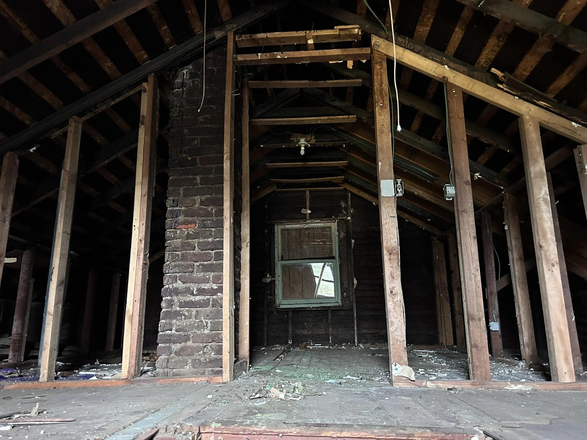 Interior of an unfinished attic with exposed wooden framing, a brick chimney, a small window, and construction debris on the floor.