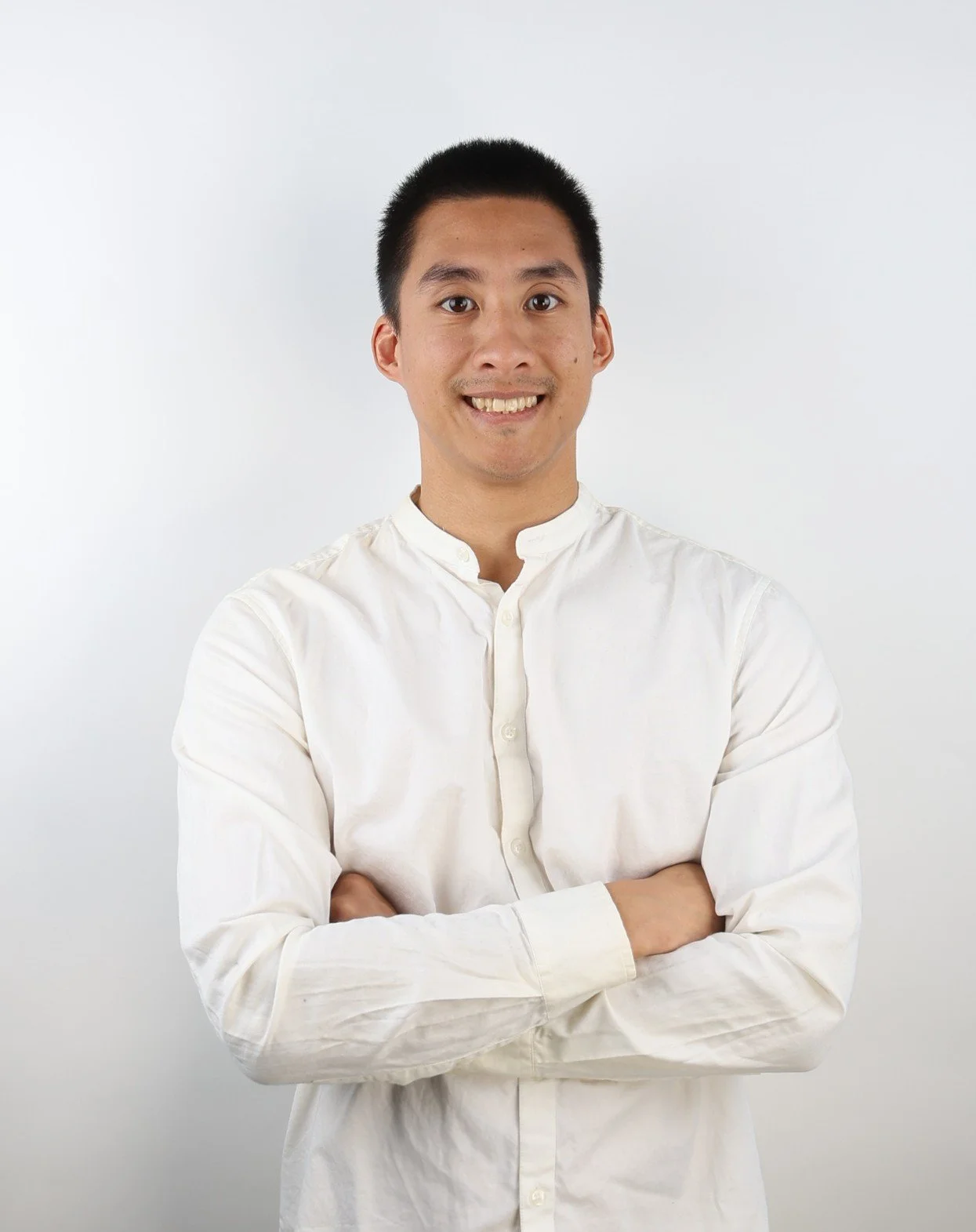 Portrait of a young man with short black hair, wearing a white button-up shirt, smiling and crossing his arms against a plain white background.