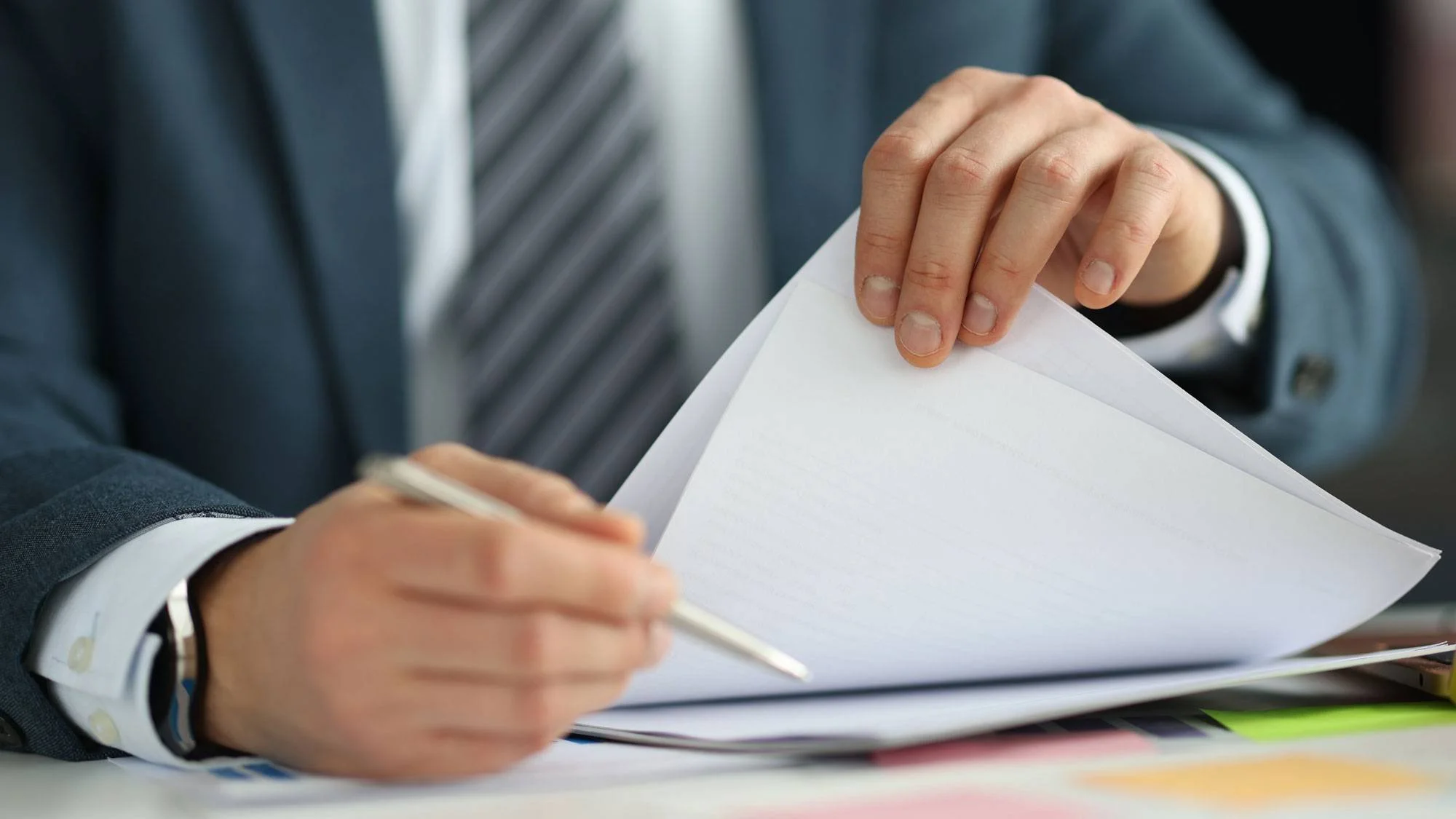 A person in a business suit appears to be reviewing or signing documents at a desk, with their hand holding a pen and flipping through papers.