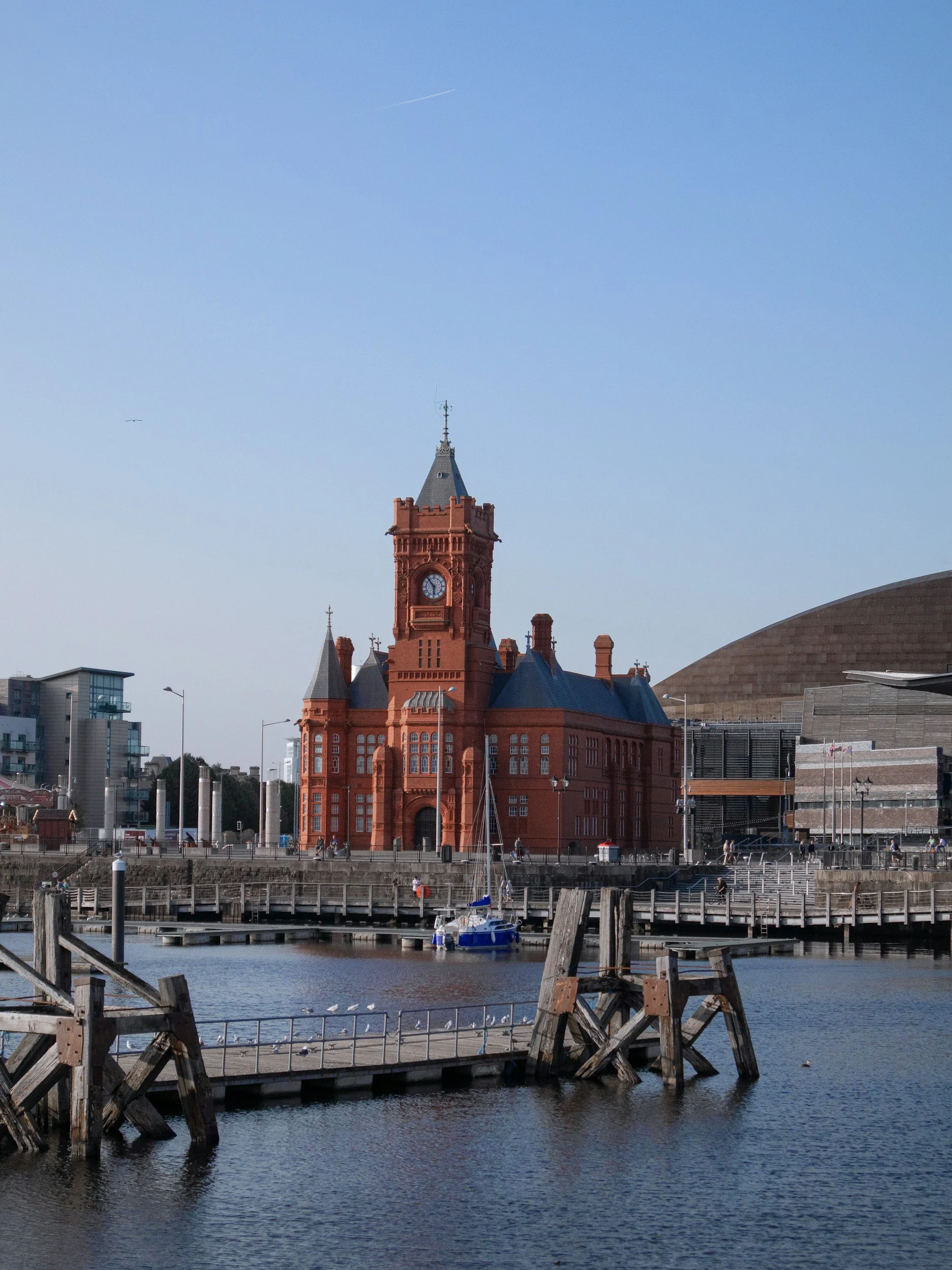 Red brick building with a clock tower near the water, sailboats, and a modern building on the right, under a clear blue sky.