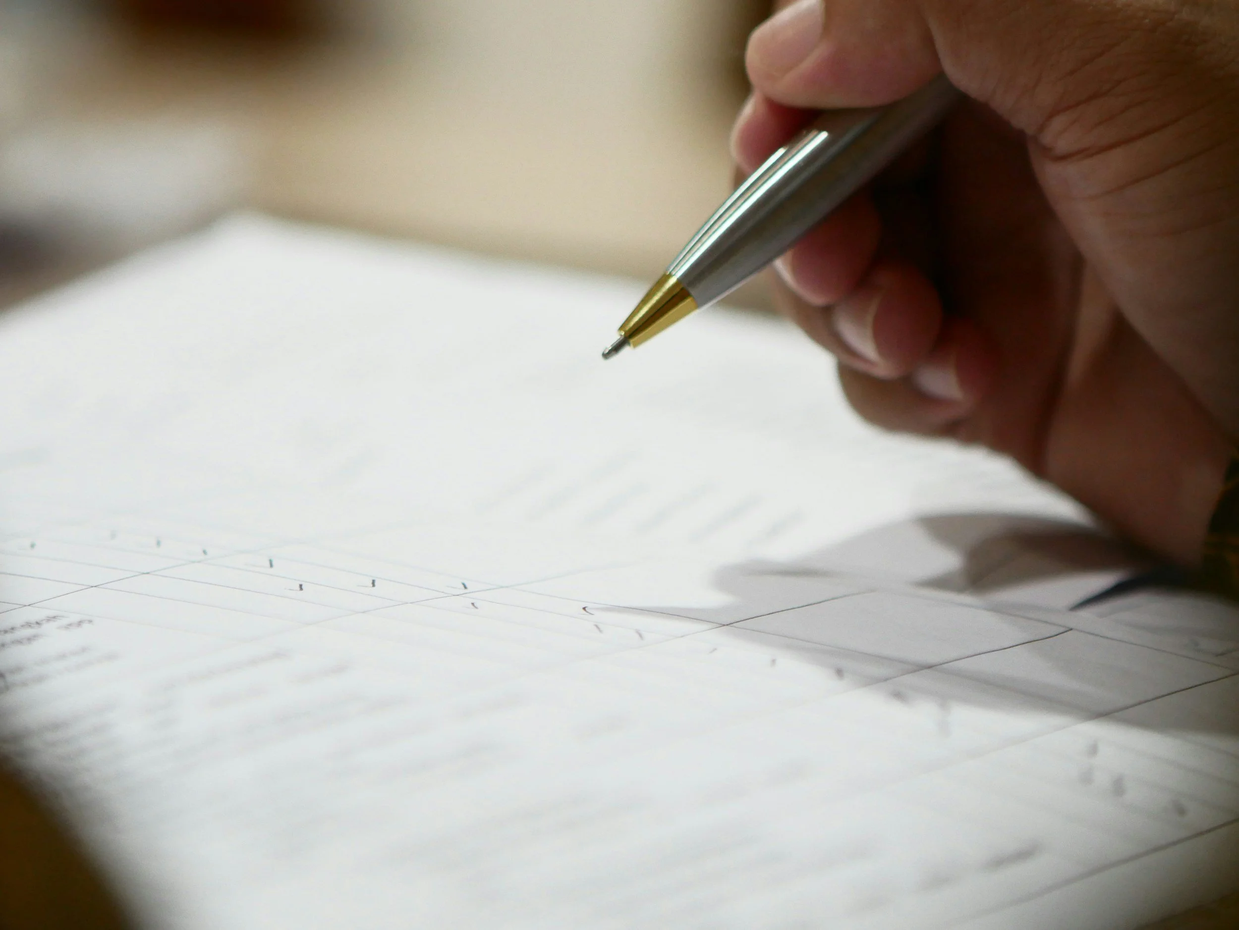 Close-up of a person's hand holding a silver and gold pen, writing on a white paper with faint lines.