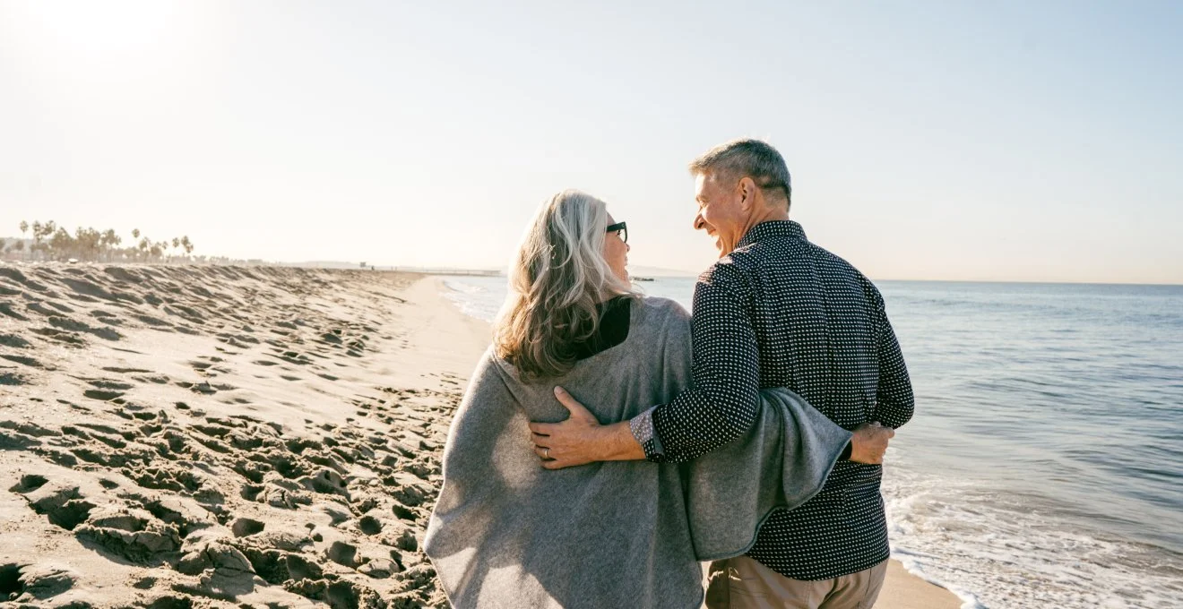 A happy couple walking on the beach, smiling and embracing each other with the ocean and clear sky in the background.