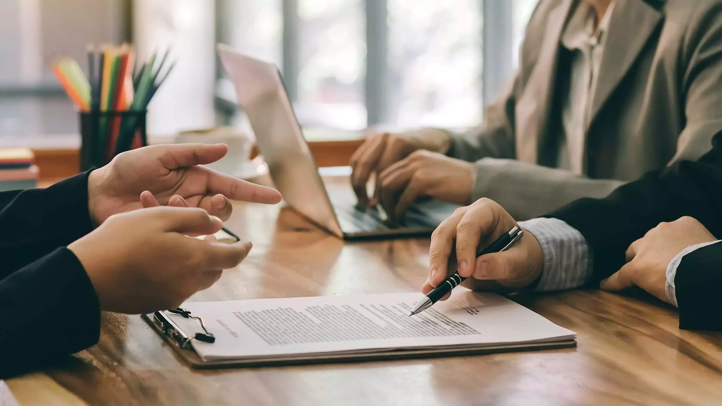 Two people sitting at a wooden table during a discussion, one is pointing at a document with a pen, and the other is typing on a laptop, with office supplies in the background.