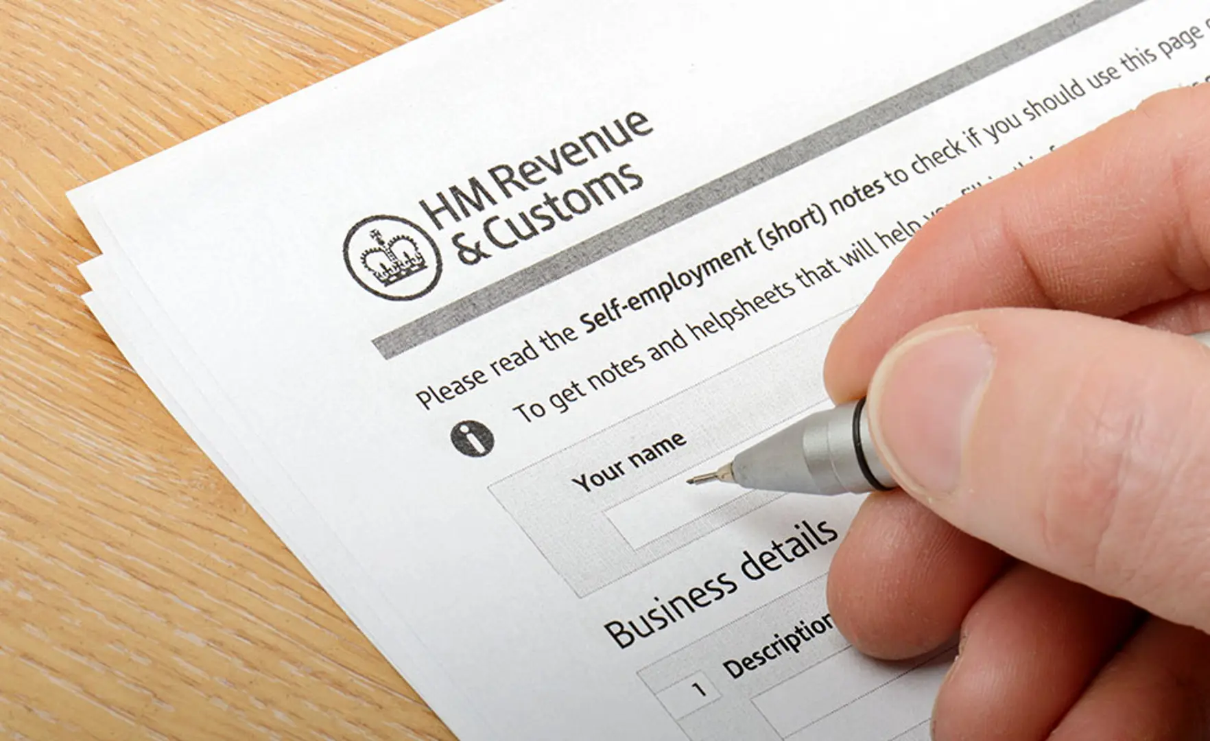 A person filling out a form from HM Revenue & Customs for self-employment, using a pen, on a wooden surface.