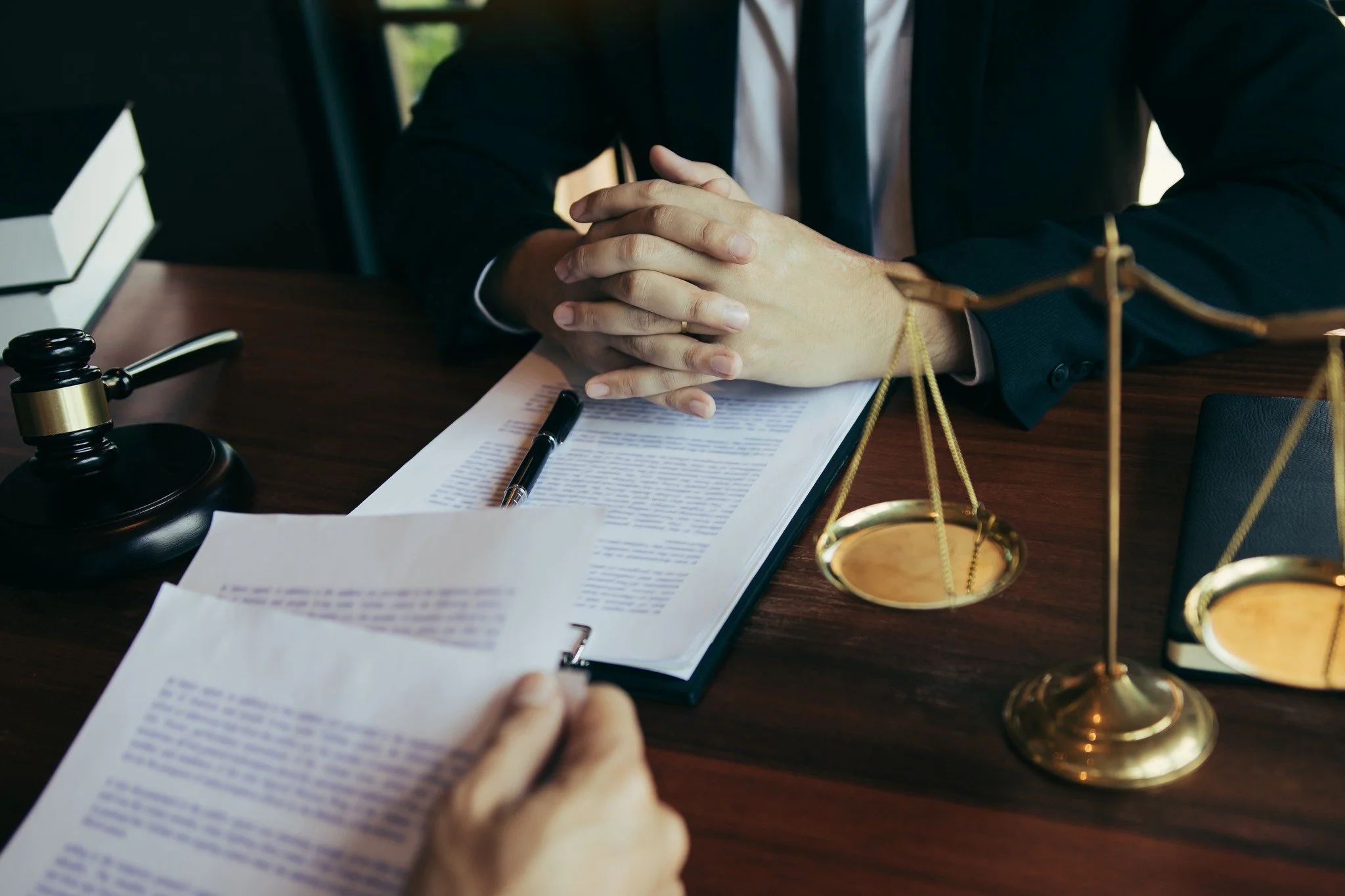A person wearing a suit with hands clasped on a desk near legal documents, a gavel, and a scale of justice.