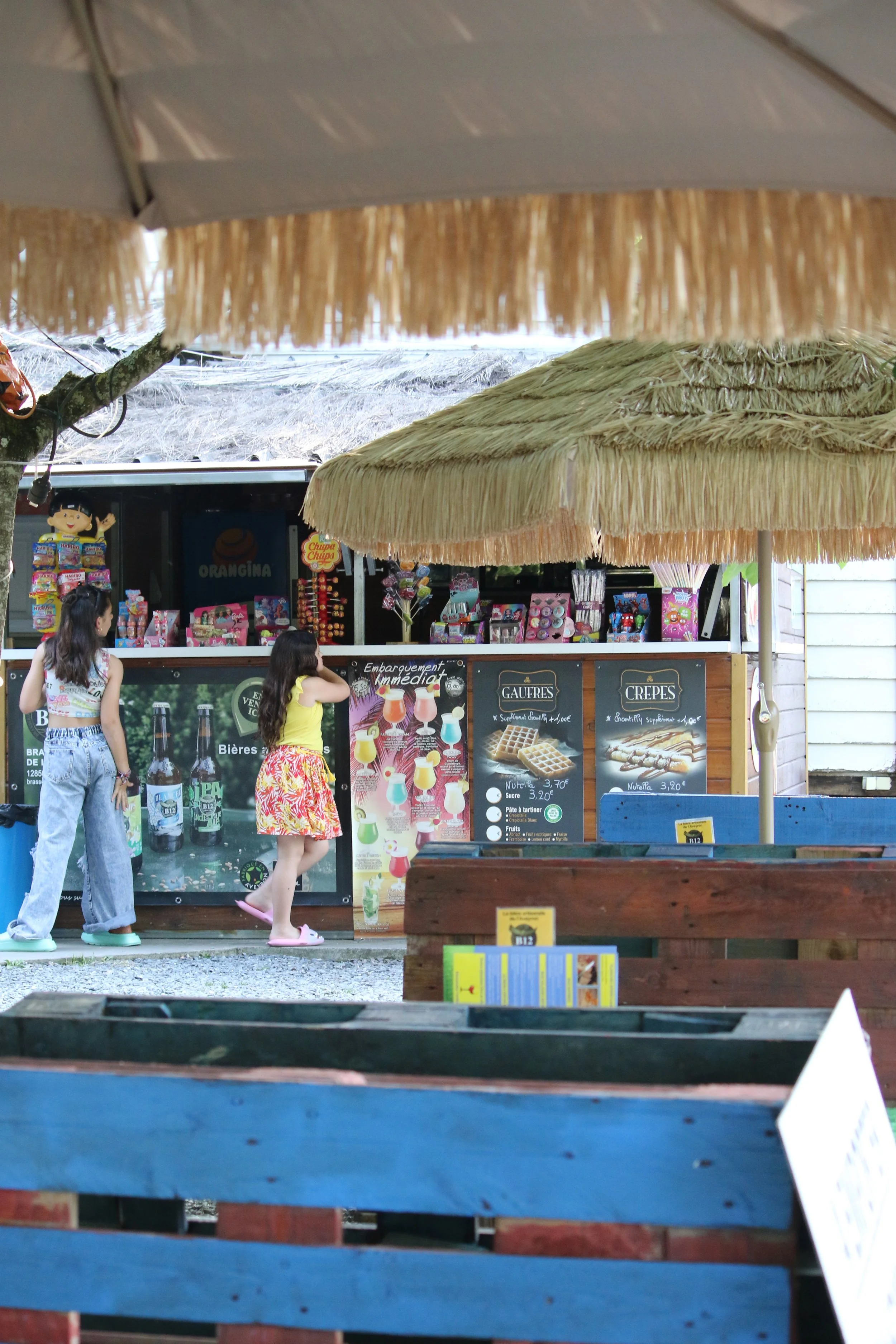 Deux enfants font la queue devant un stand de nourriture ou de boissons dans un lieu en extérieur, avec des parasols en paille au-dessus.