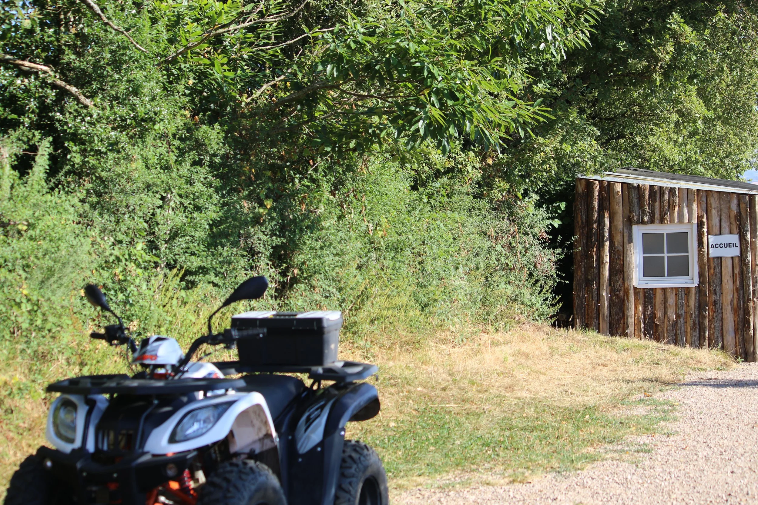 Une petite cabane en bois avec une fenêtre et une porte portant une plaque 'ACCUEIL', entourée de végétation dense, avec un quad passant à proximité.