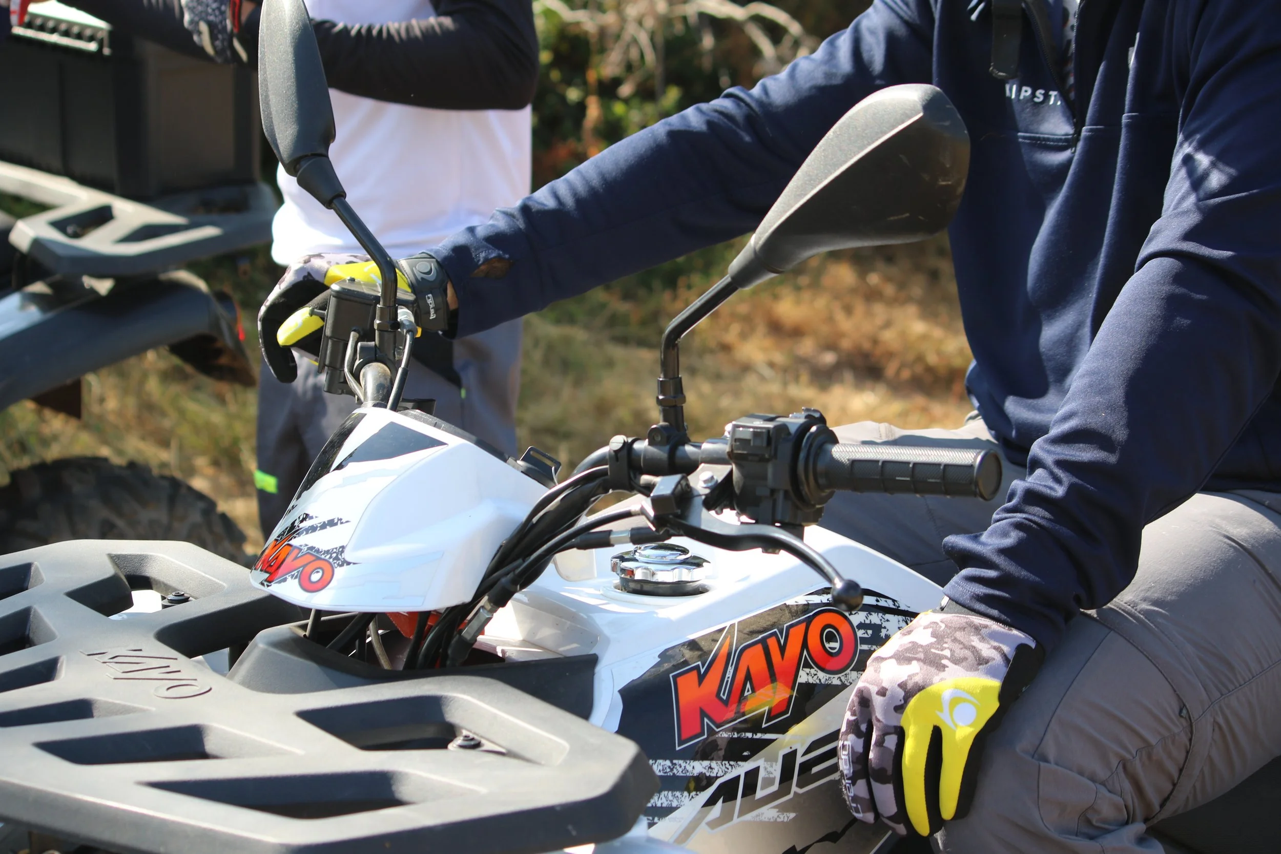 Personne montant une moto tout-terrain White KAYO, portant des gants jaunes et un haut bleu, avec d'autres personnes en arrière-plan.