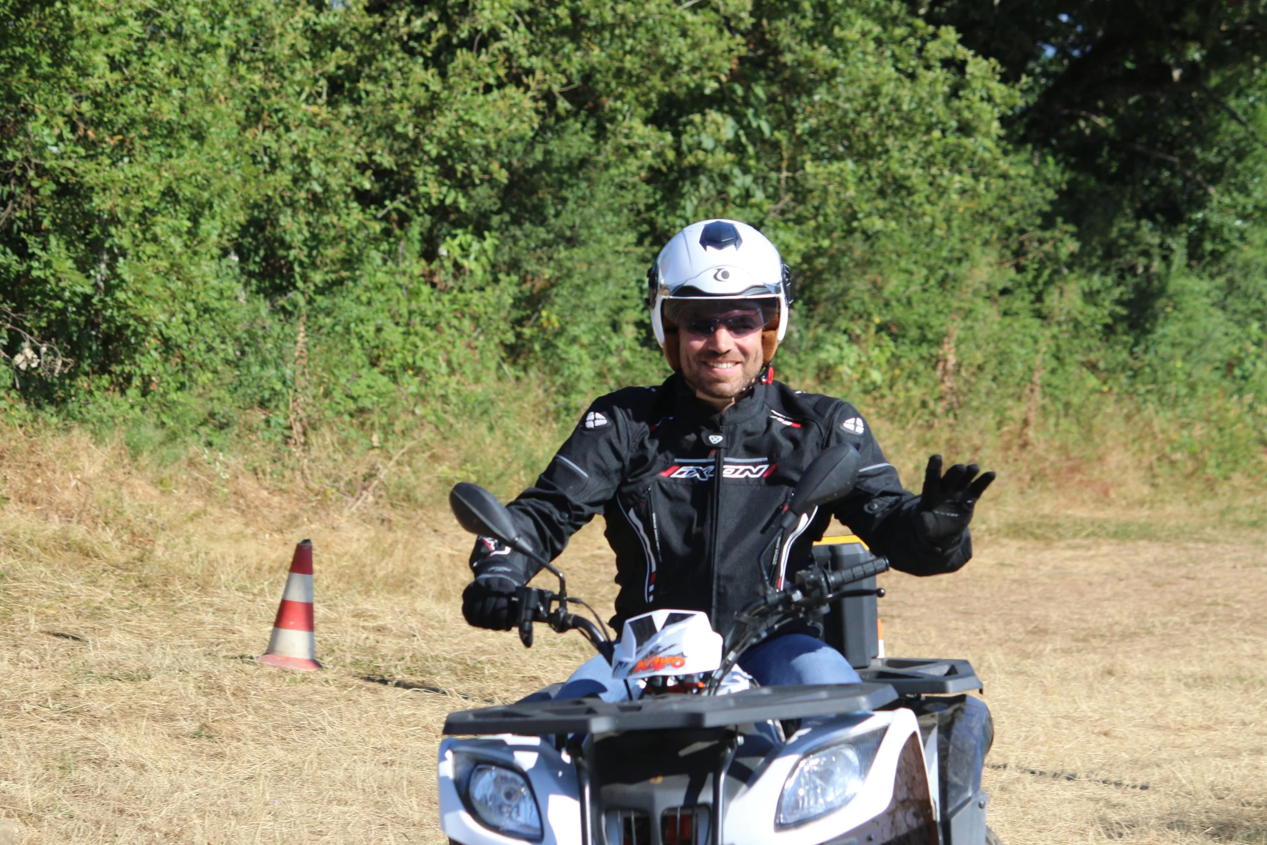 Homme en tenues de moto et casque, manœuvrant un quad dans un espace extérieur herbeux, avec un cône orange- blanc en arrière-plan.