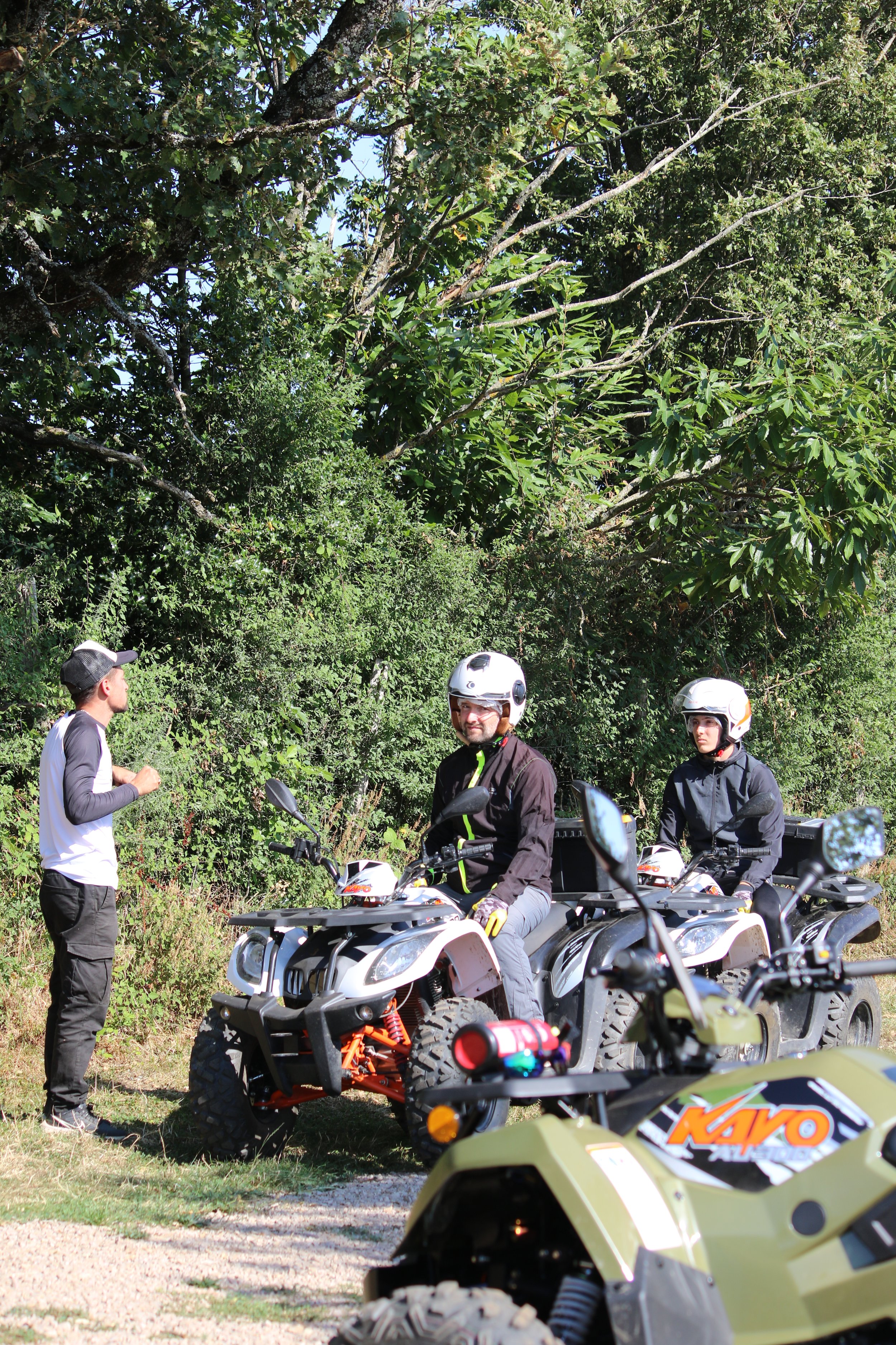 Trois personnes en tenues de motarde avec casques, deux sur des quad et une autre debout à côté, dans un environnement naturel avec des arbres.