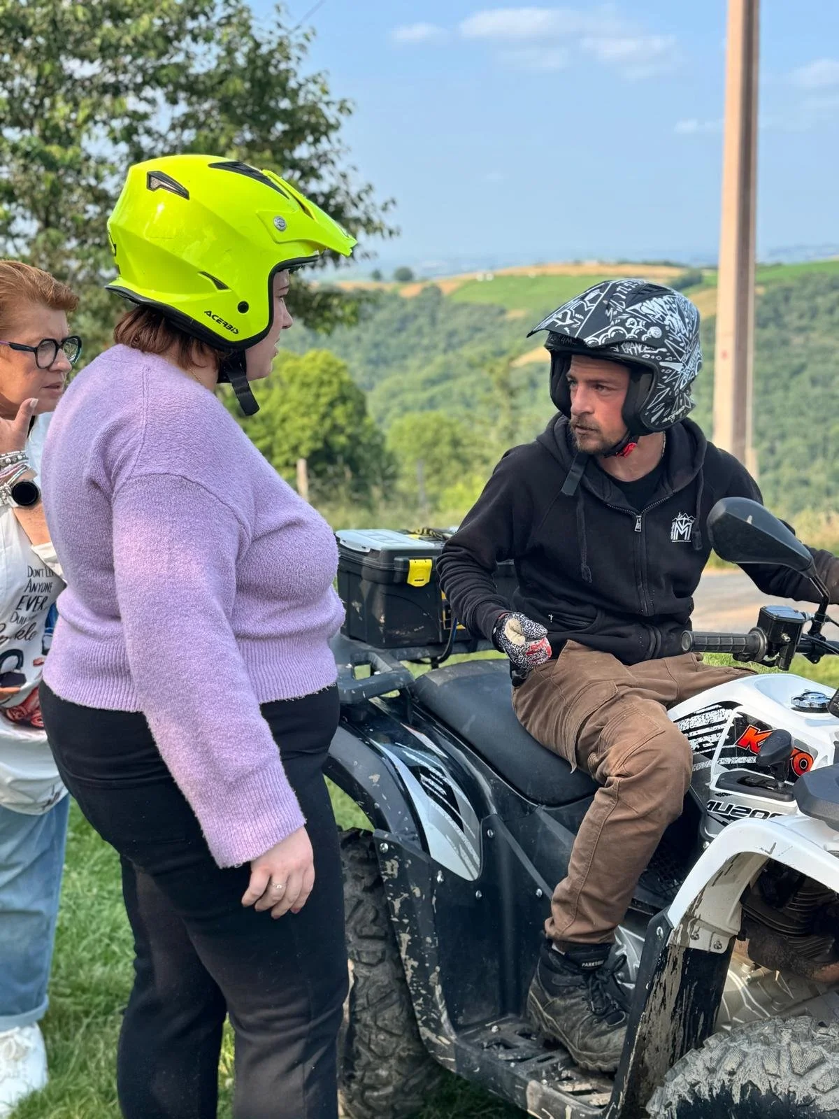 Trois personnes, dont une à moto tout-terrain, discutent en extérieur avec un champ et des collines en arrière-plan, deux d'entre elles portent un casque.