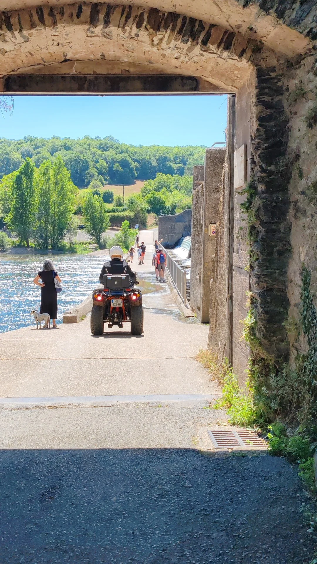 Vue d'un chemin ou trottoir le long d'une rivière, avec des personnes, une femme avec un chien, une moto, un groupe de marcheurs, et un paysage verdoyant en arrière-plan, vu à travers une arche en pierre.