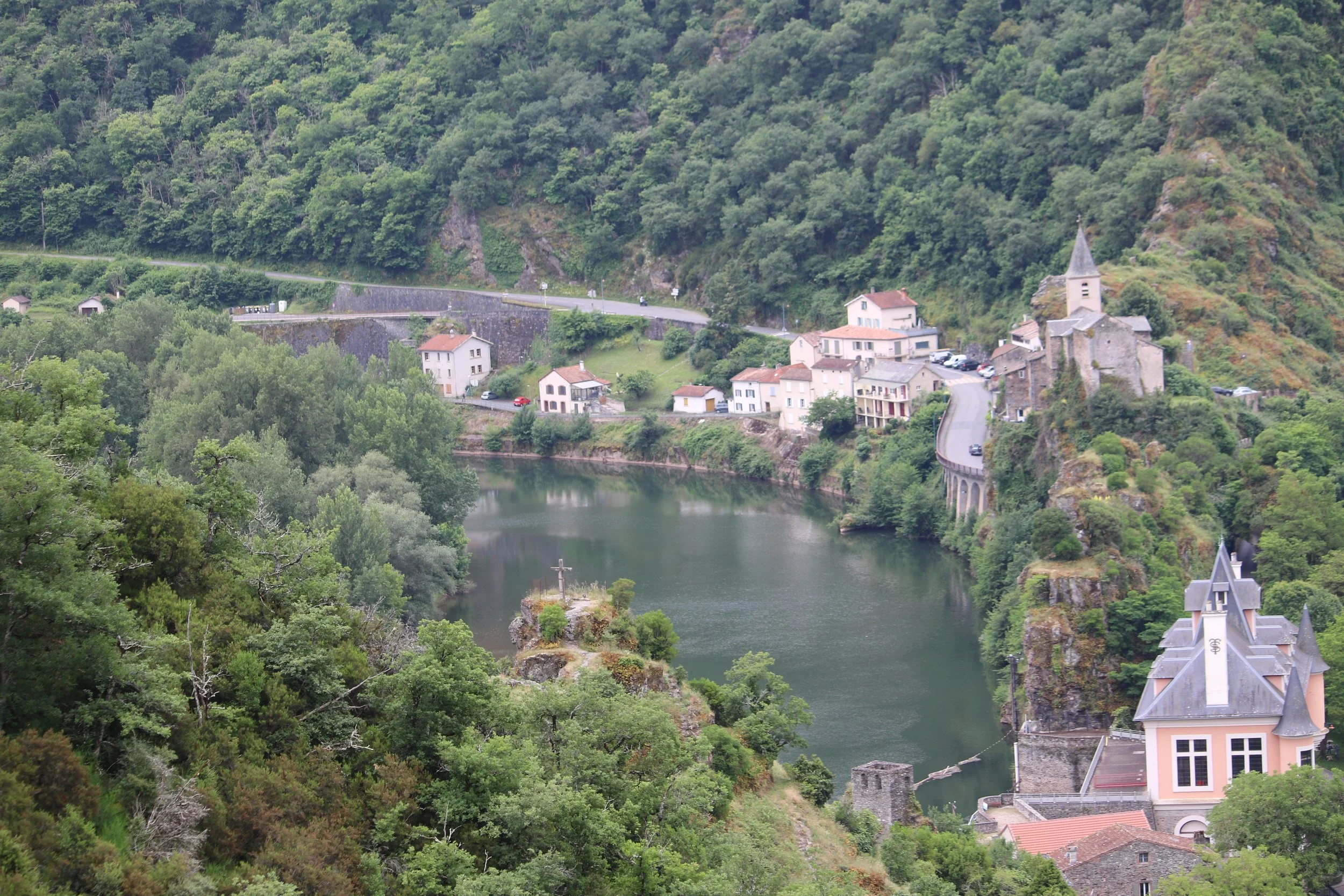 Un village au bord d'une rivière entouré de collines verdoyantes avec des maisons, une église avec un clocher, et un petit pont en pierre.